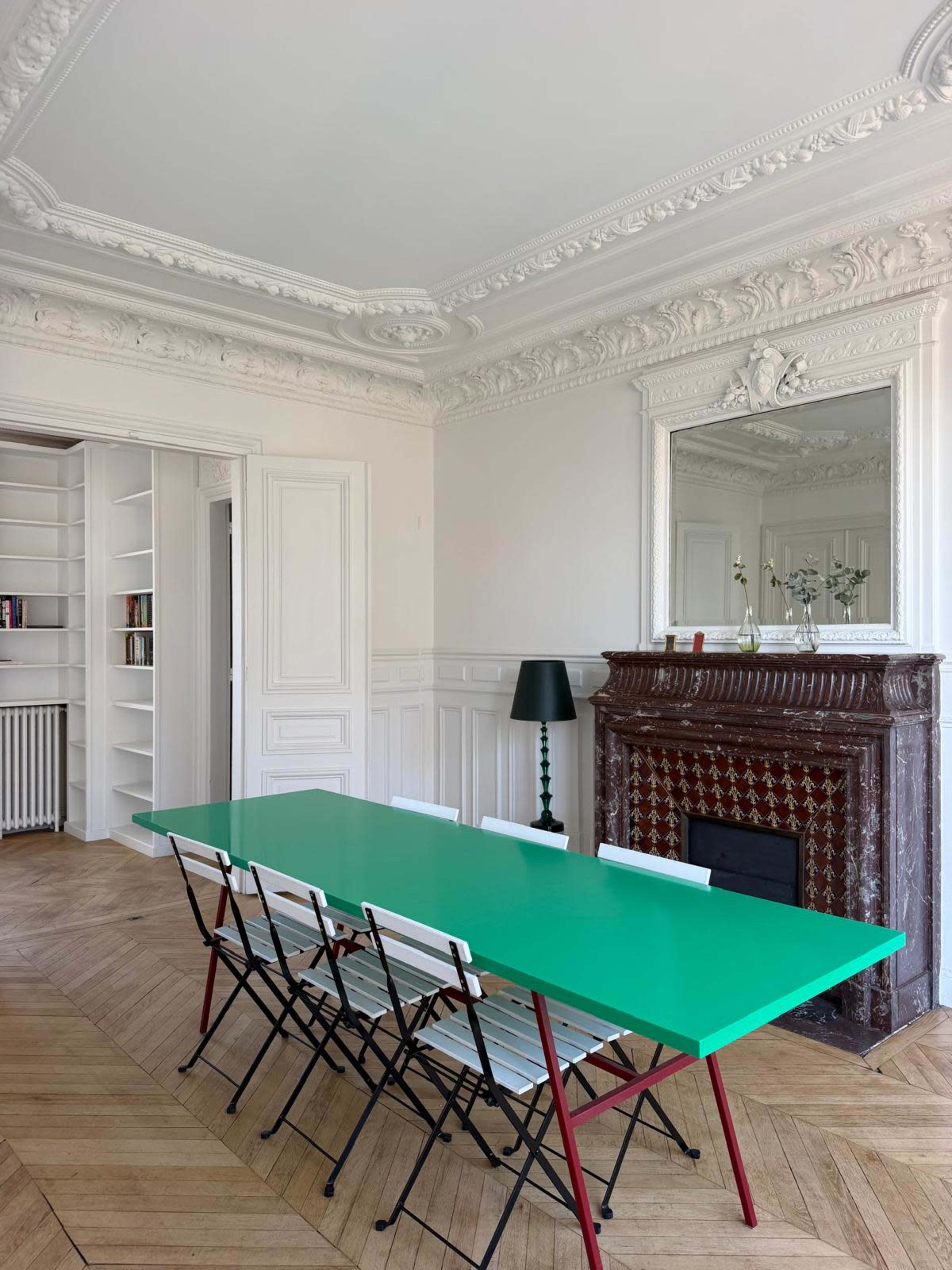 The image shows a minimalist dining area with a bright green table surrounded by black and white folding chairs, a decorative fireplace, and a large mirror reflecting the room's architectural details.