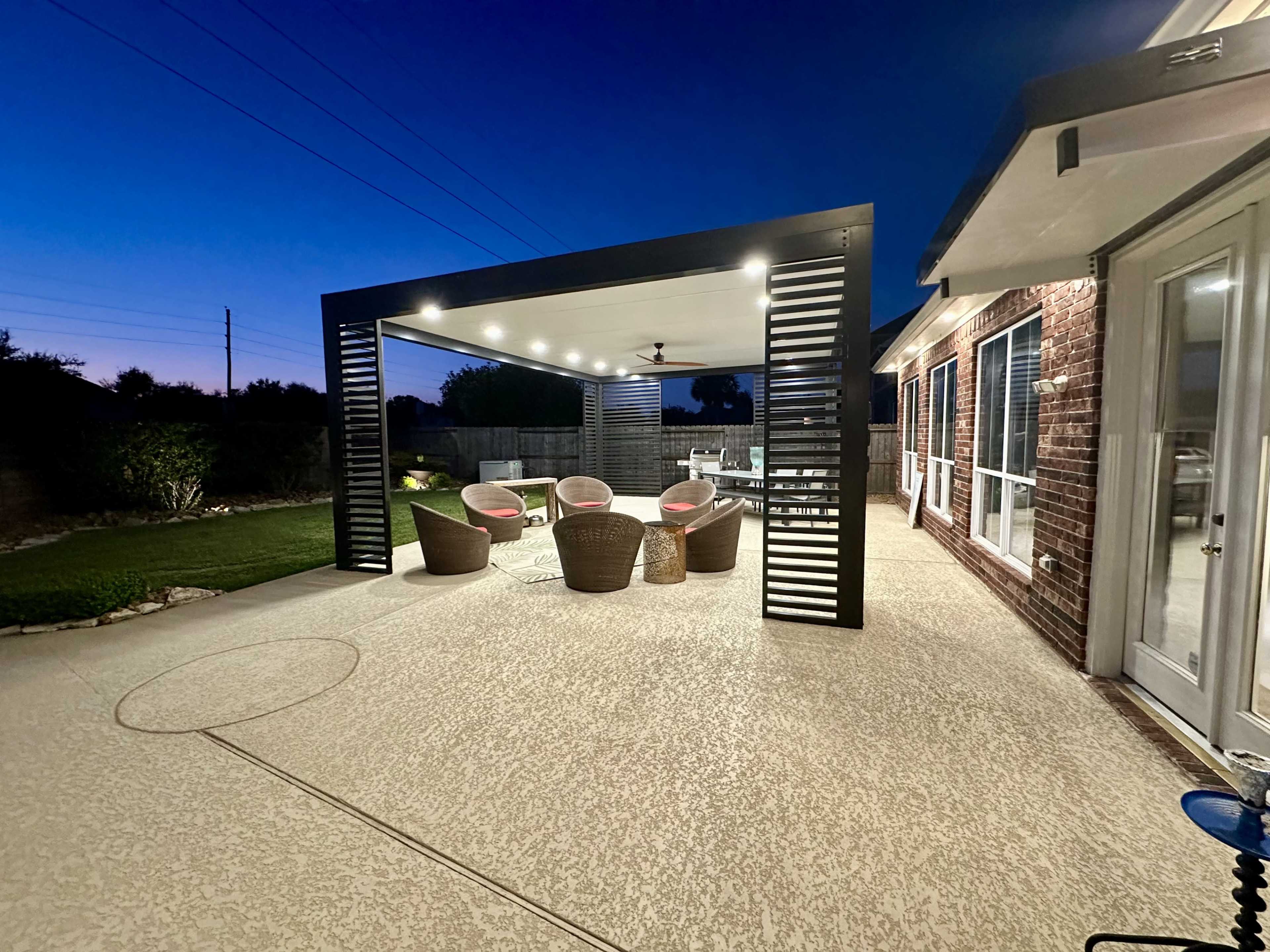 A covered outdoor patio features four wicker chairs and a table, illuminated under a darkening sky.