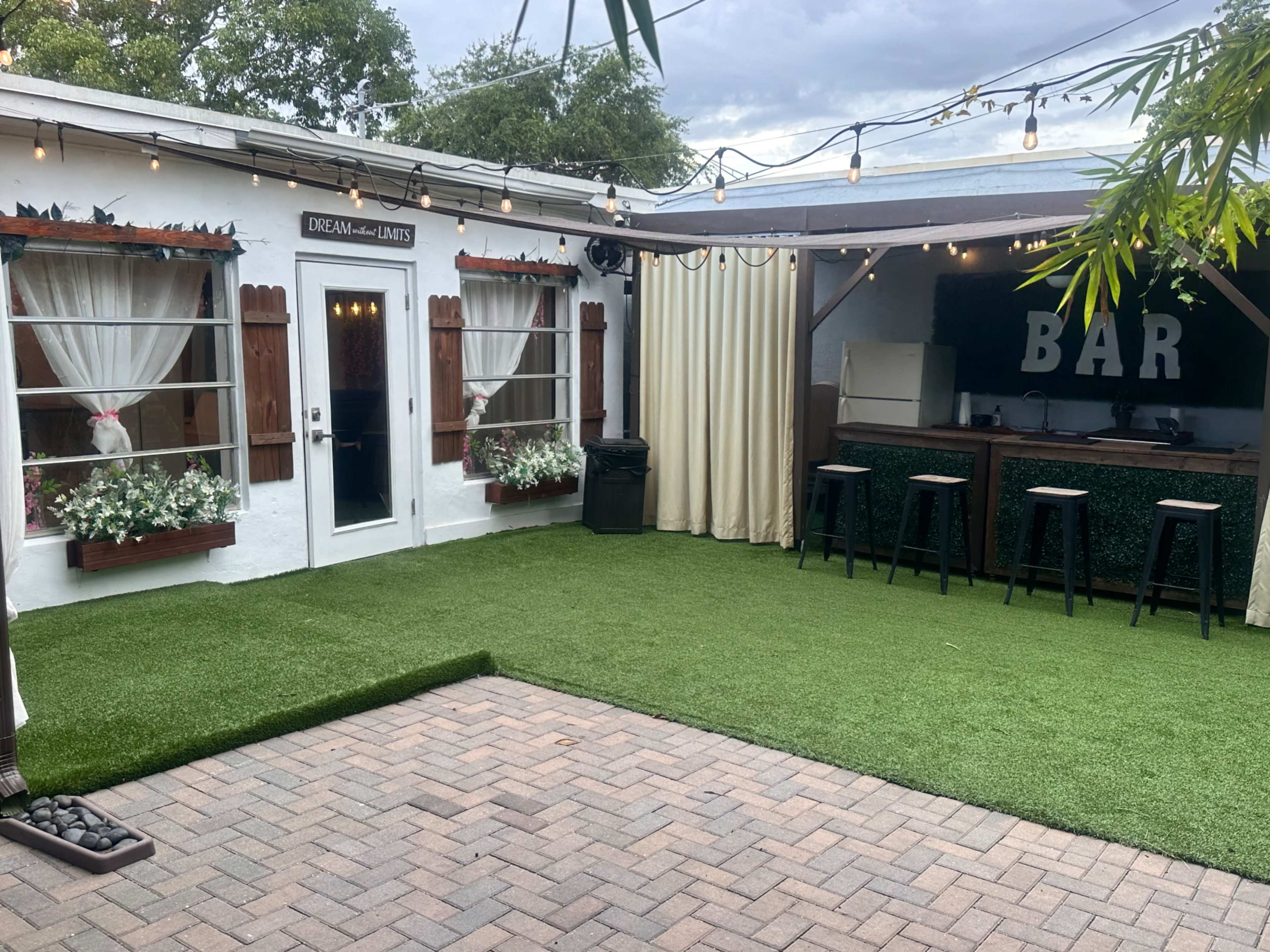 The image shows an outdoor space featuring a bar with stools, surrounded by greenery and decorative curtains, on a paved area.