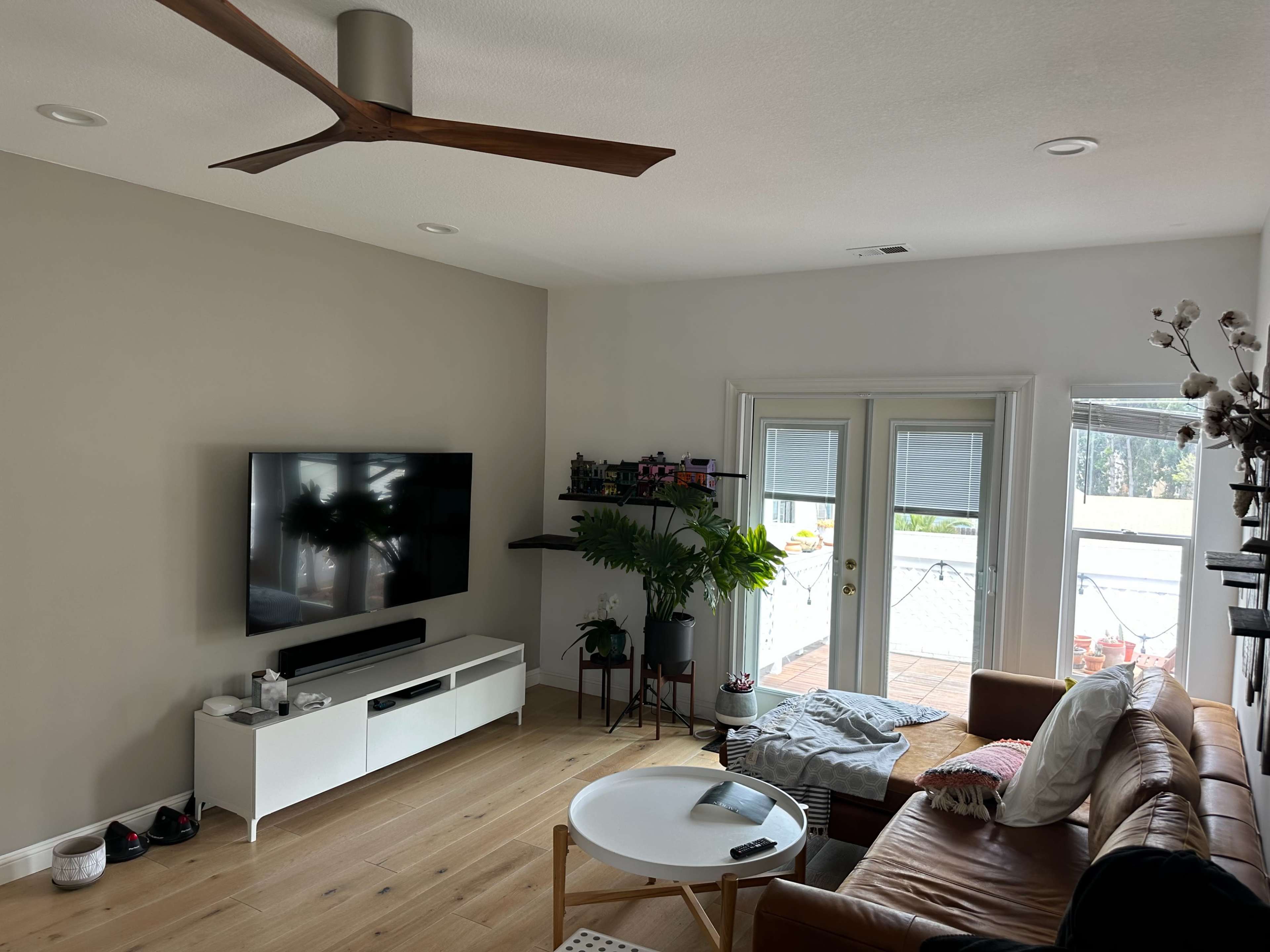 A spacious living room featuring a modern TV unit, a brown leather sofa, a round coffee table, and large windows with a view of an outdoor area.