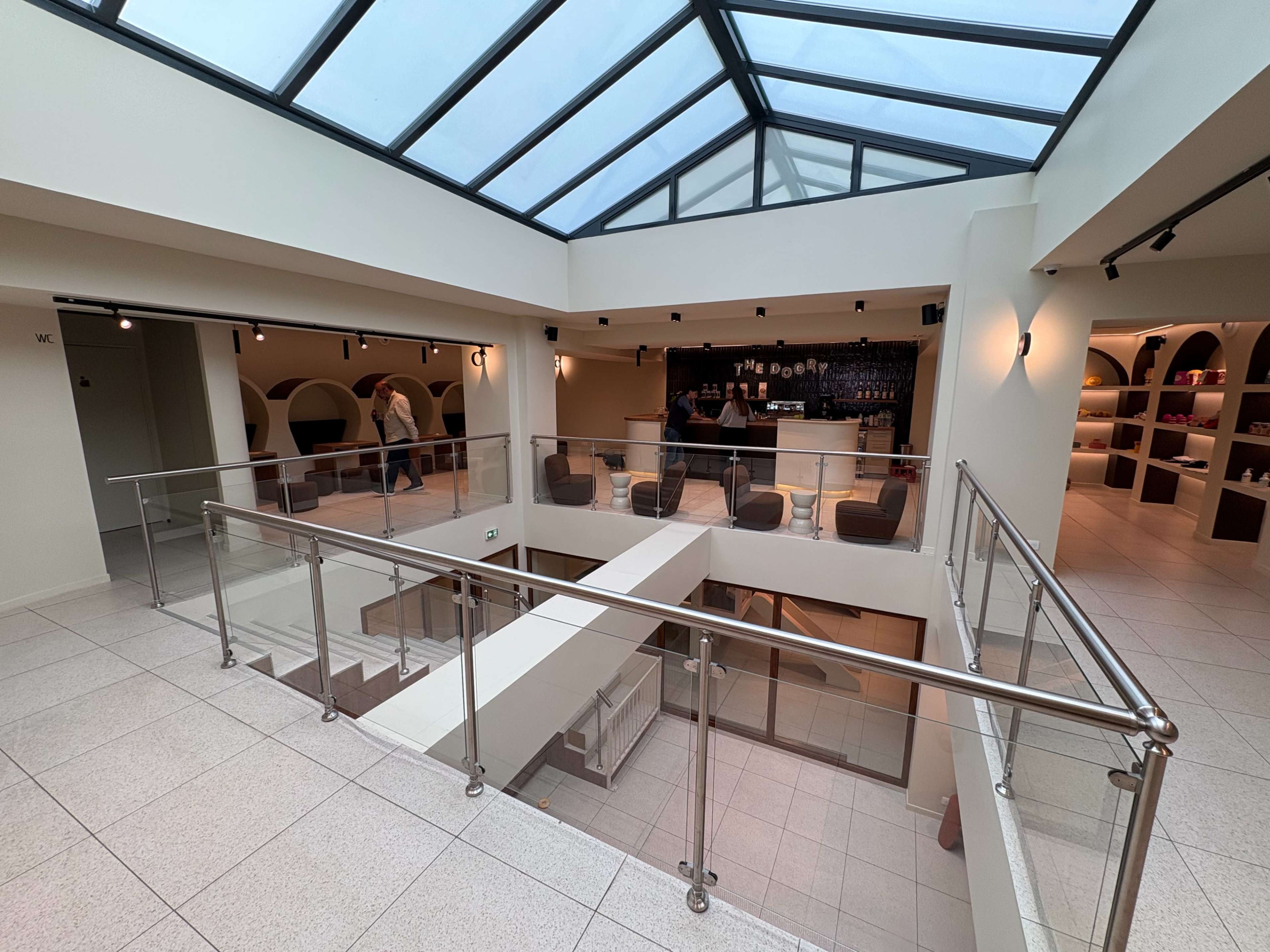 The image shows a modern interior space with a glass ceiling, featuring a balcony overlooking a lower level with seating and a bar area.