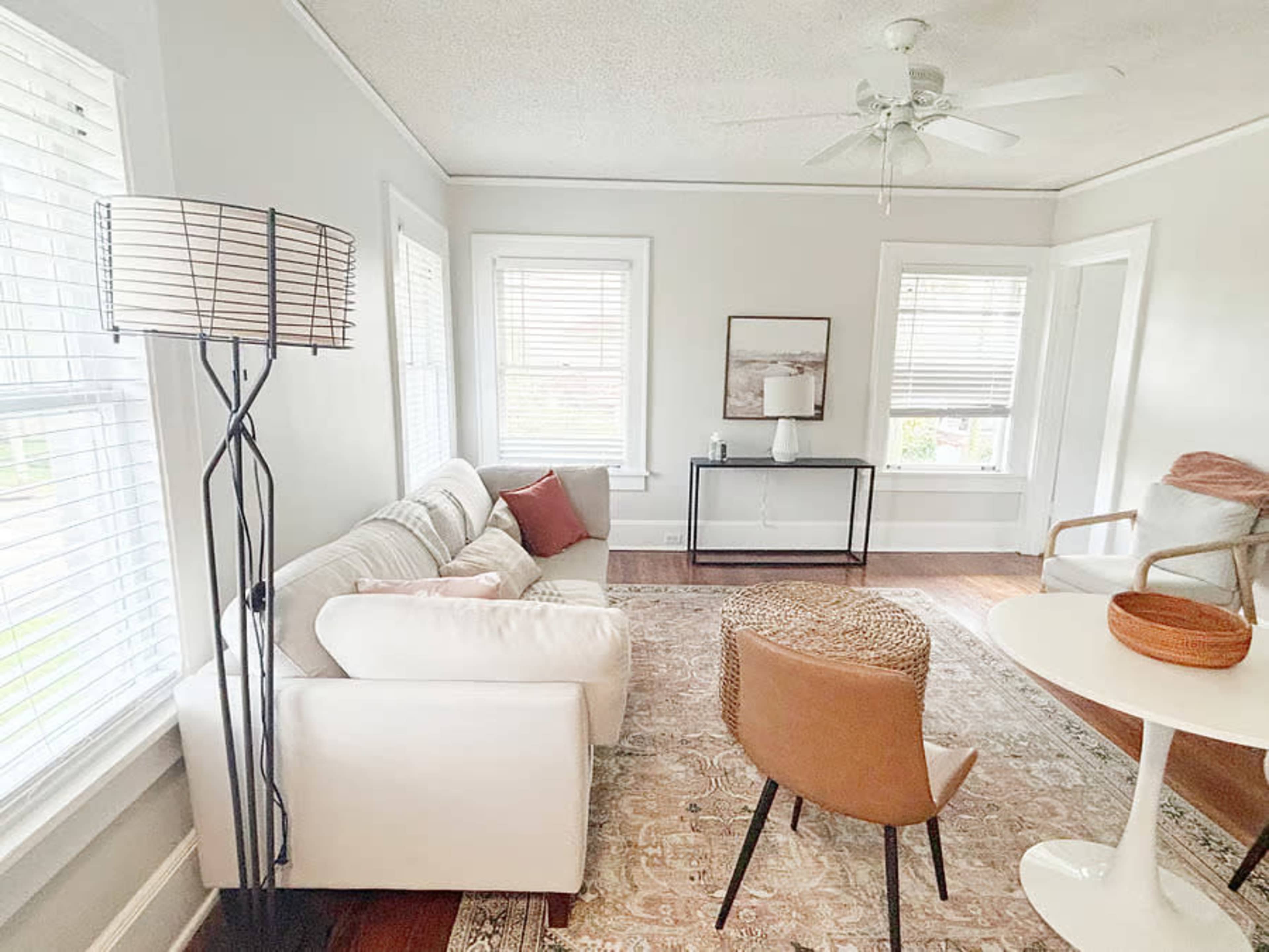 The image shows a bright living room with a light gray color scheme, featuring a white sofa, a round table, and a tall lamp beside a window.
