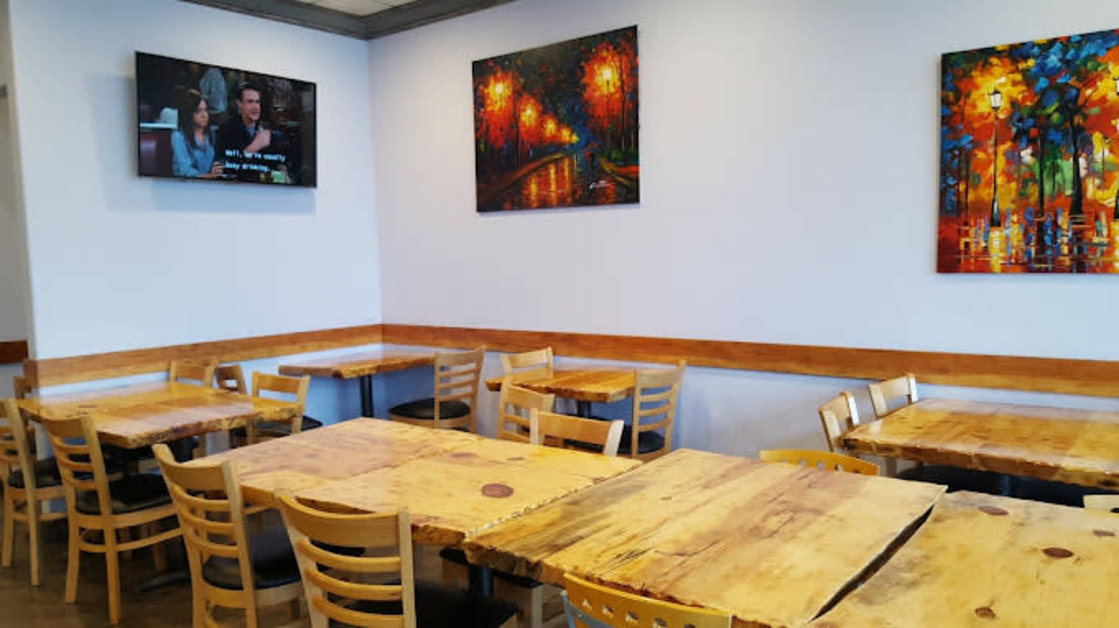 The image shows a restaurant interior featuring wooden tables and chairs, with colorful artwork on the walls and a television displaying a program.
