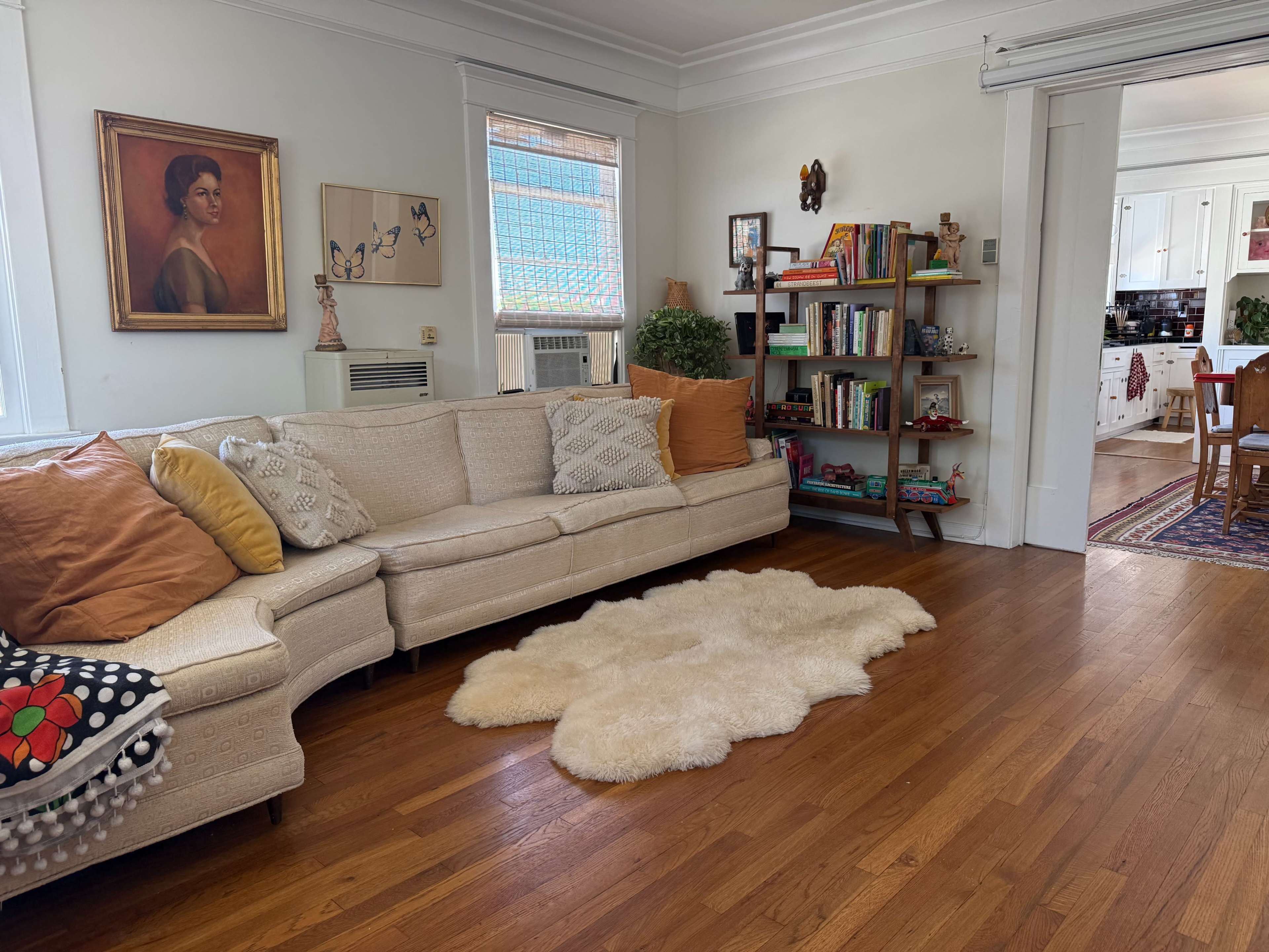 The image shows a cozy living room with a beige sectional sofa adorned with pillows, a fluffy white area rug, a bookshelf filled with books, and an adjacent kitchen area visible through an open door.