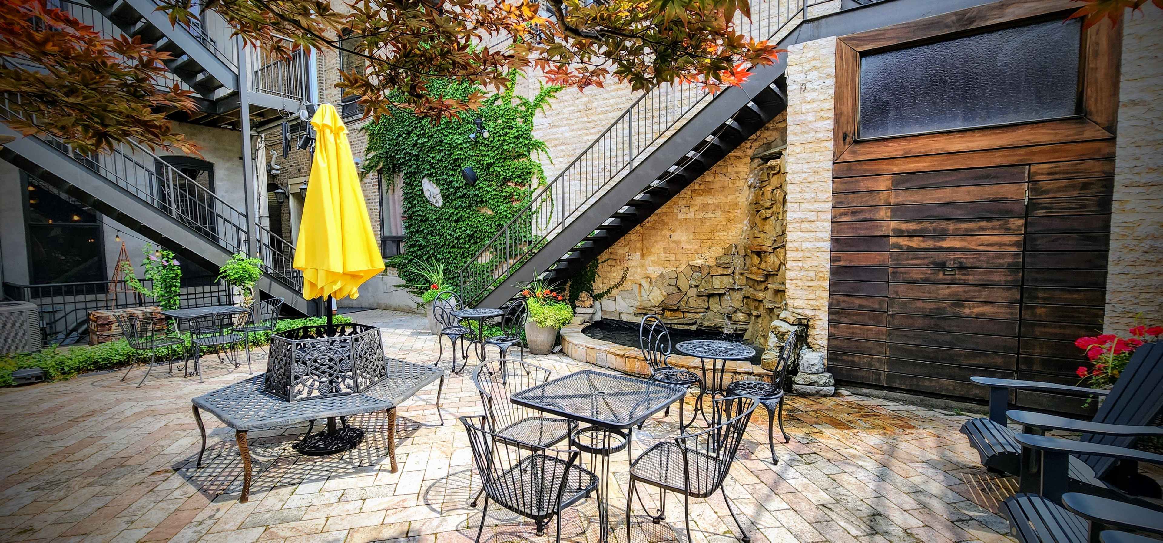 A courtyard features metal tables and chairs surrounding a small fountain, with a bright yellow umbrella providing shade beside a stone wall.