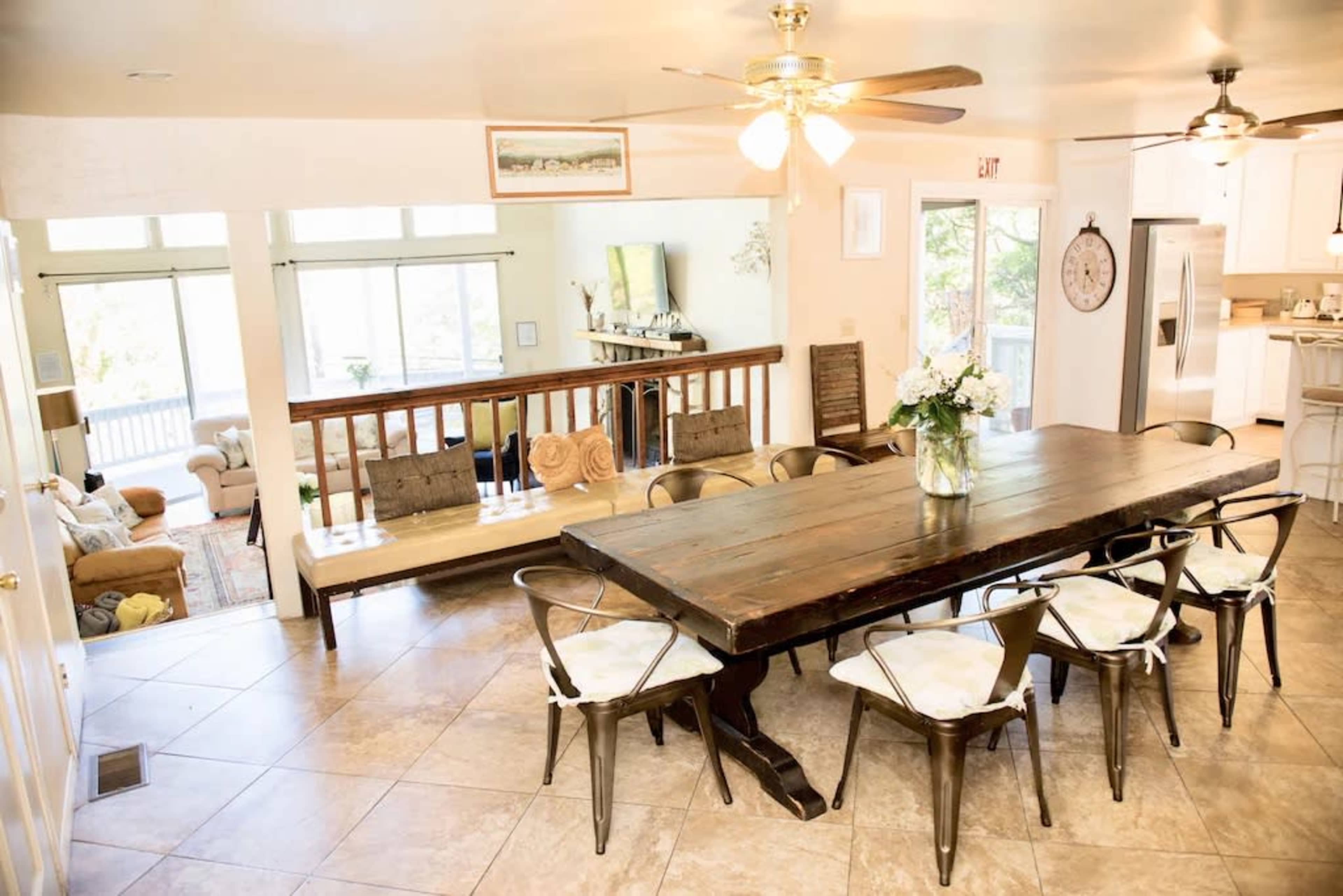The image shows a spacious dining area with a large wooden table surrounded by metal chairs, opening up to a brightly lit living space in the background.