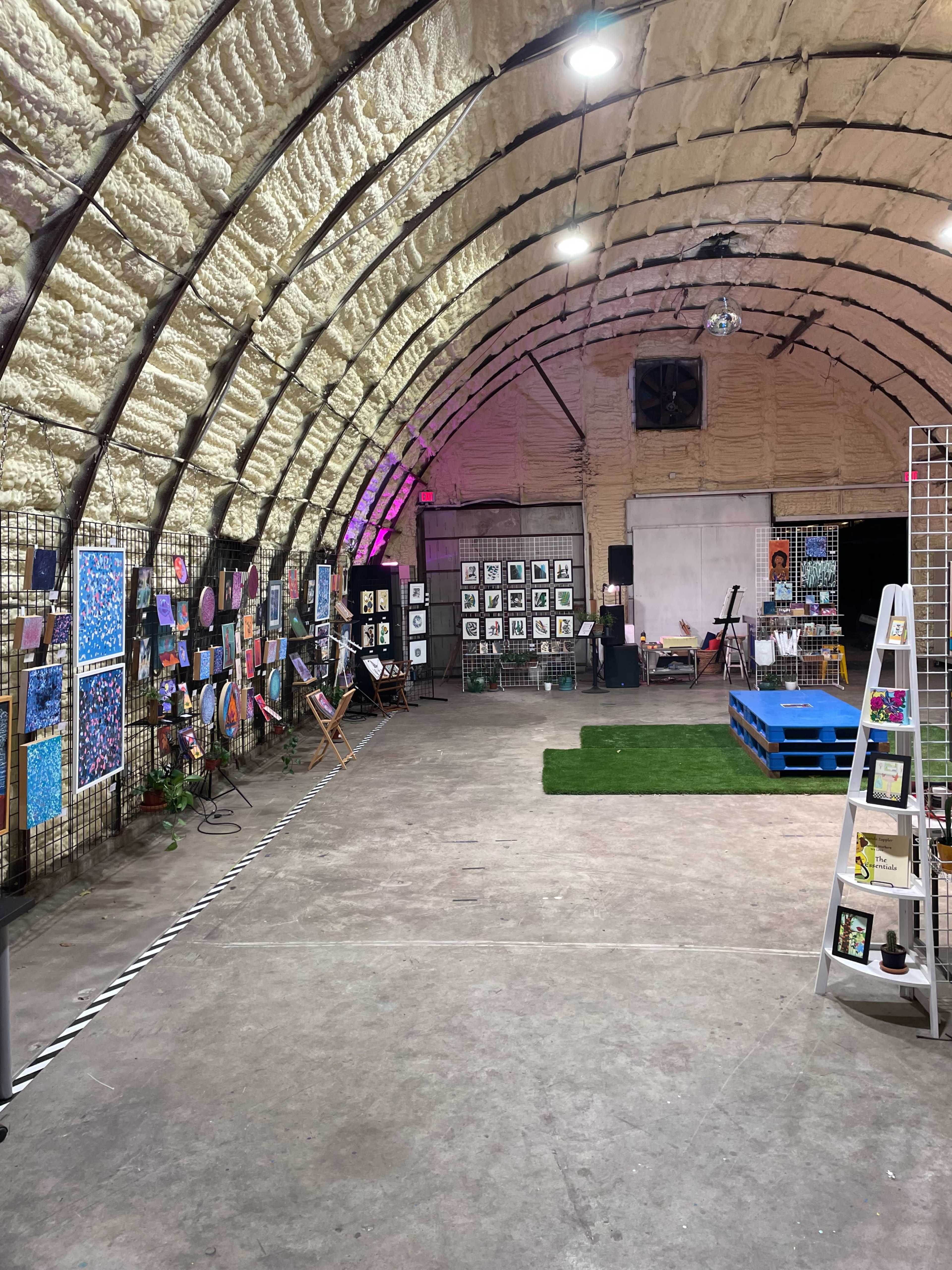 An indoor art exhibit featuring various paintings and artworks displayed on walls with a curved ceiling, set in a spacious, industrial-style environment.