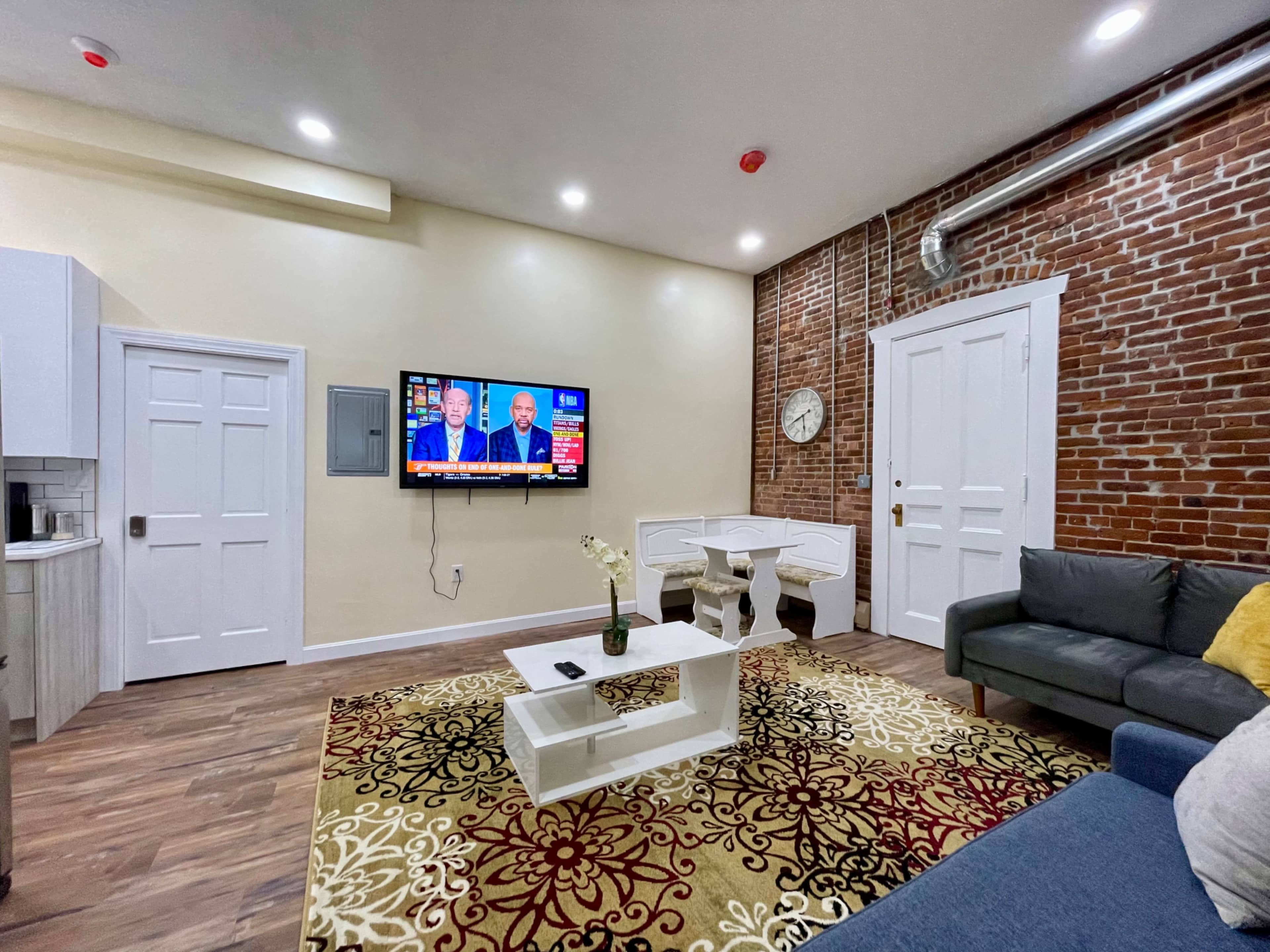 The living area features a television mounted on a wall with a brick backdrop, a gray sofa, a white coffee table, and a patterned area rug.
