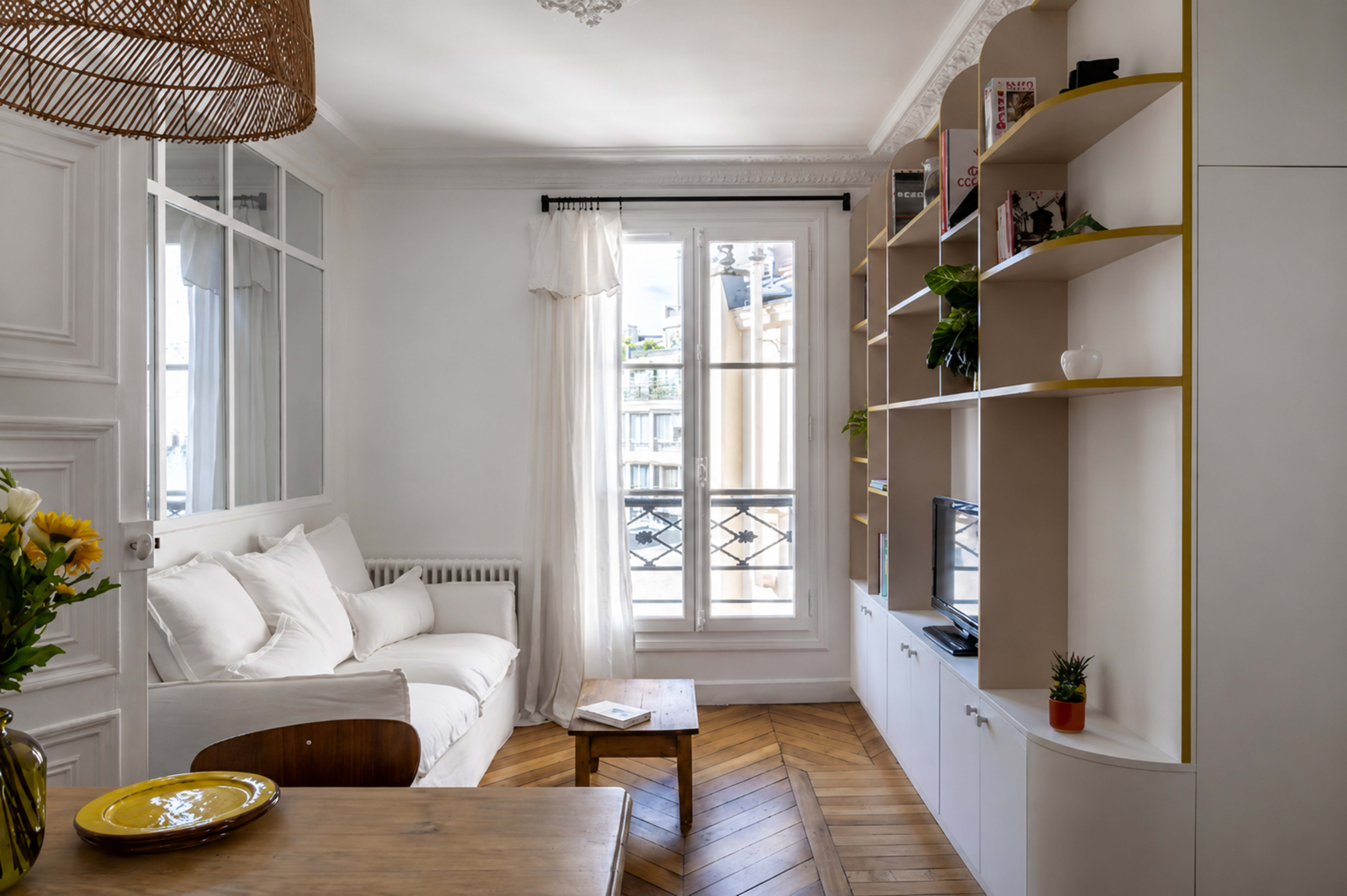 The image shows a bright, compact living room featuring a white sofa, a wooden dining table, and a wall-mounted bookshelf filled with plants and decorative items.