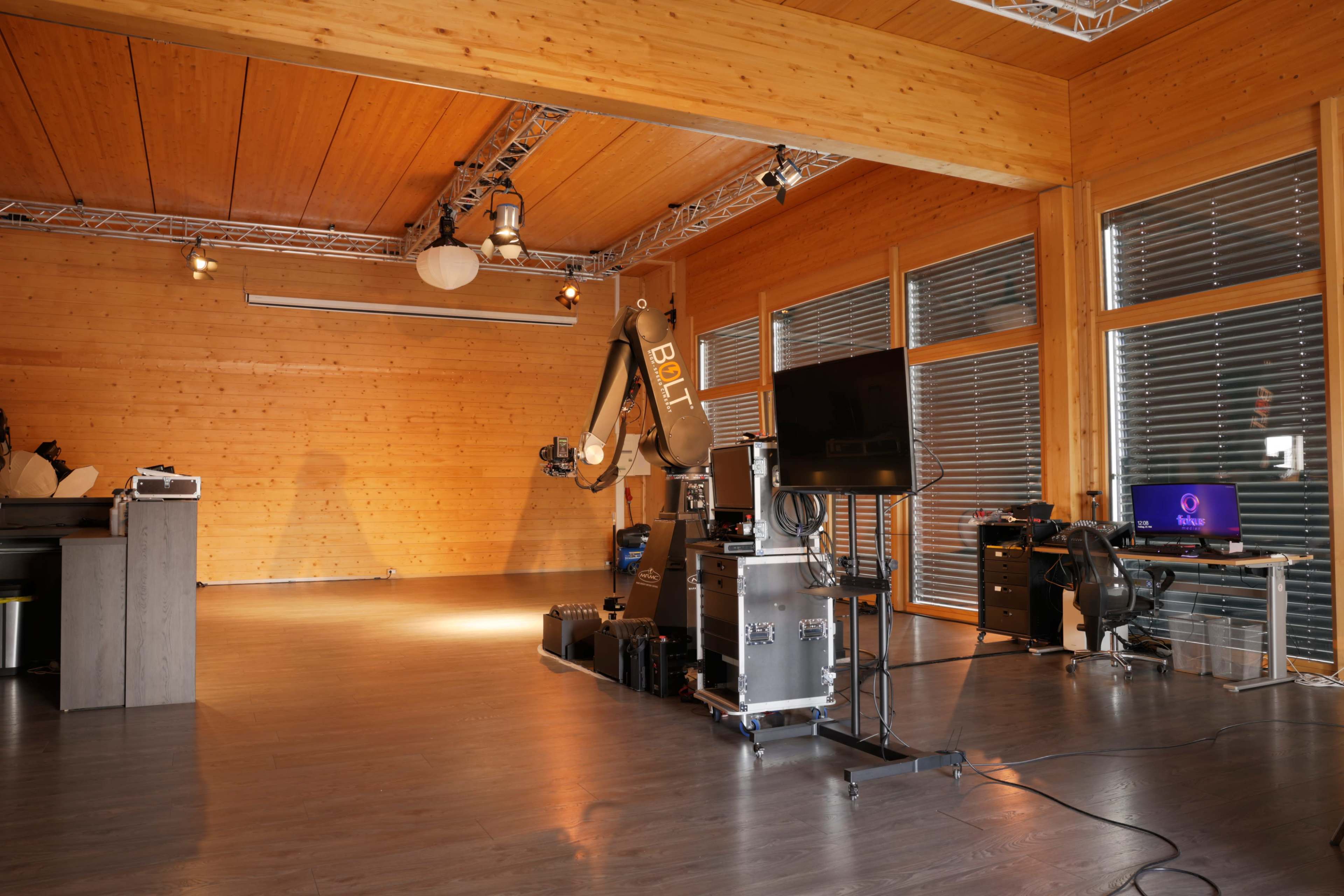 The image shows a spacious room with wooden walls and a ceiling, featuring a robotic arm, a monitor, and various equipment arranged on the floor.