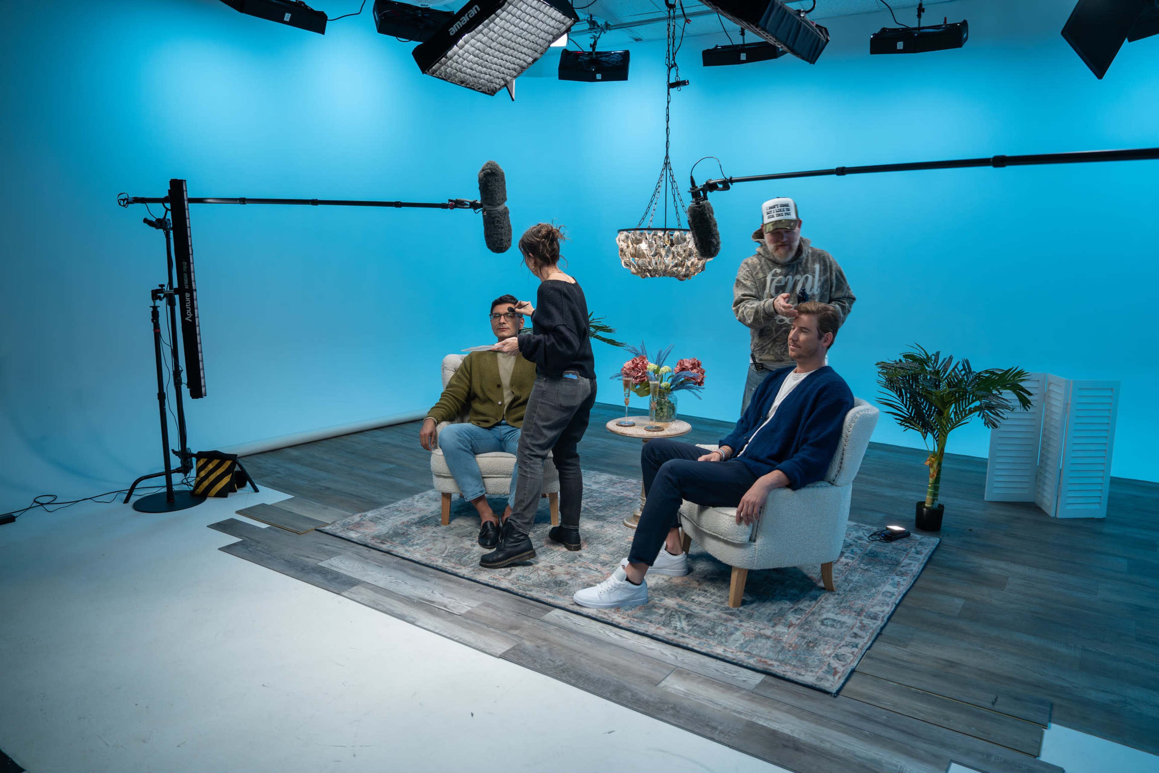 A group of four people is preparing for a video shoot in a studio with a blue background and several lighting and sound equipment setups.