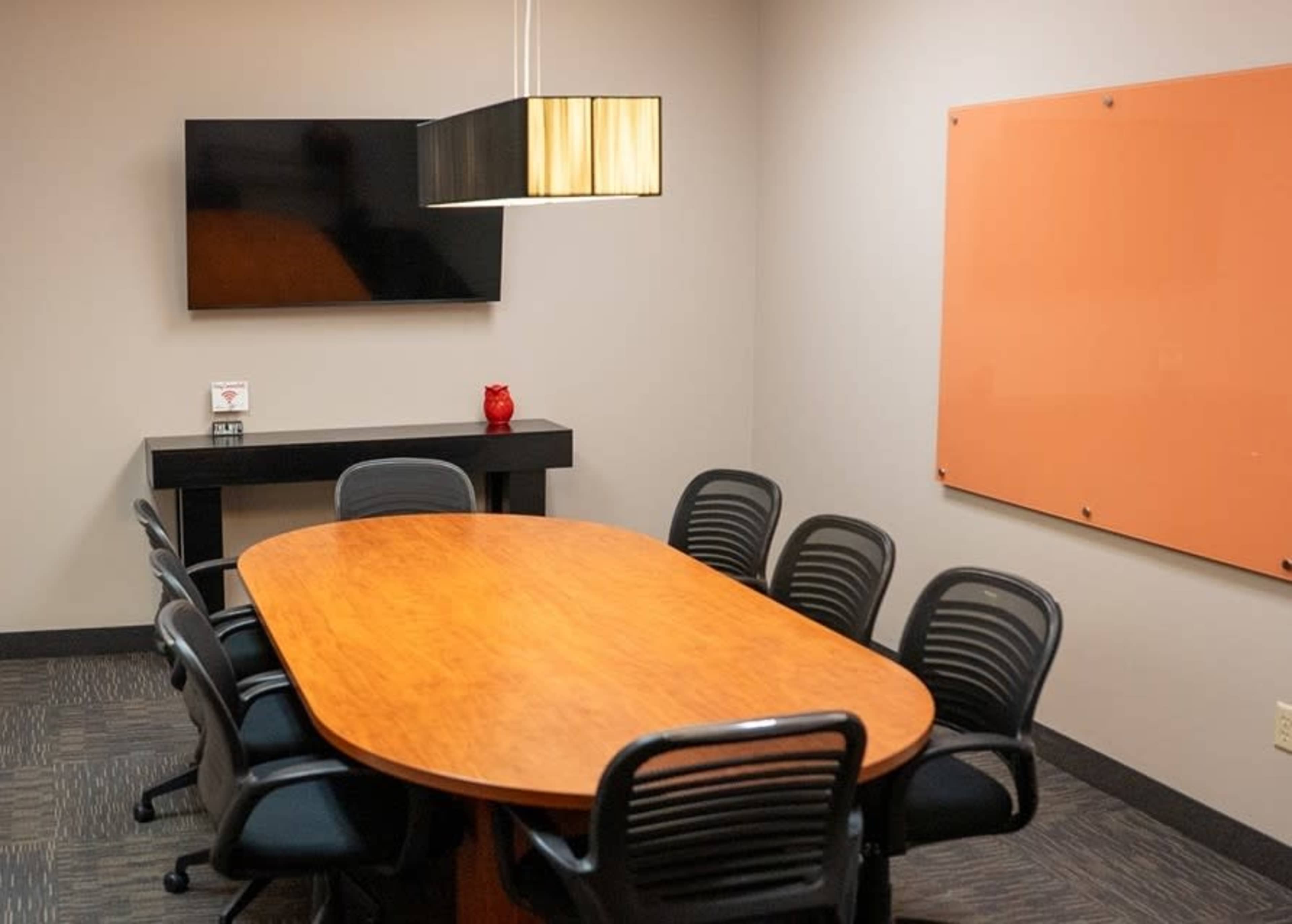 The image shows a conference room with a large oval wooden table surrounded by eight black mesh chairs, a flat-screen television mounted on the wall, and an orange whiteboard.