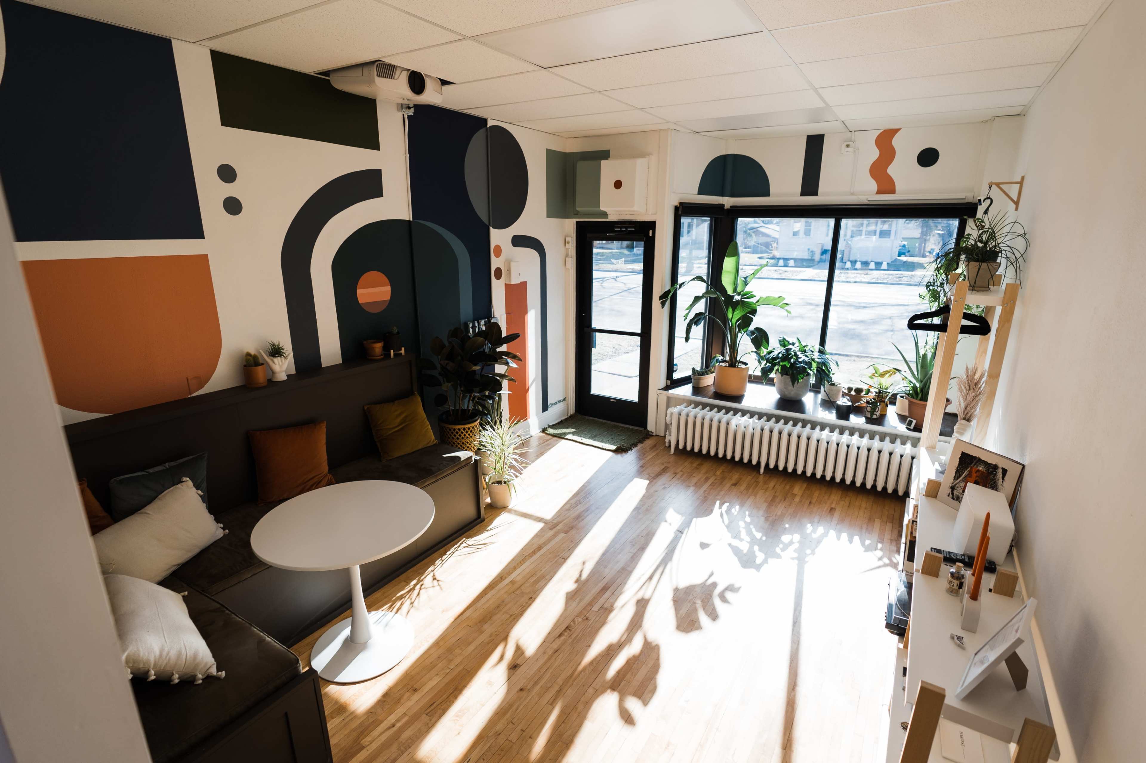 A modern, brightly lit room with a sectional sofa, a round table, and large windows displaying potted plants and geometric wall art.
