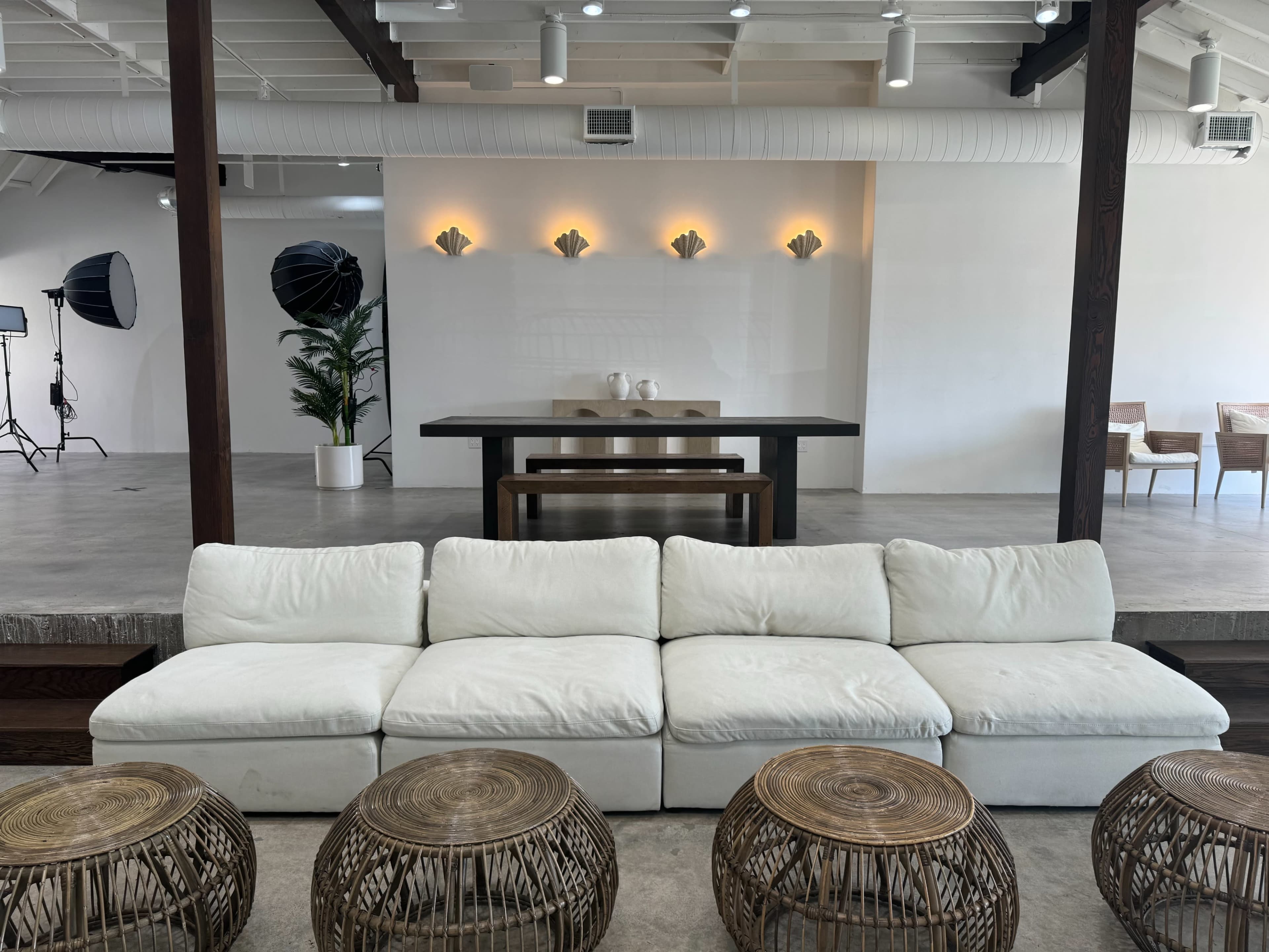 A spacious interior featuring a white sectional sofa, round wicker tables, and modern lighting fixtures on the wall behind a central dining table.