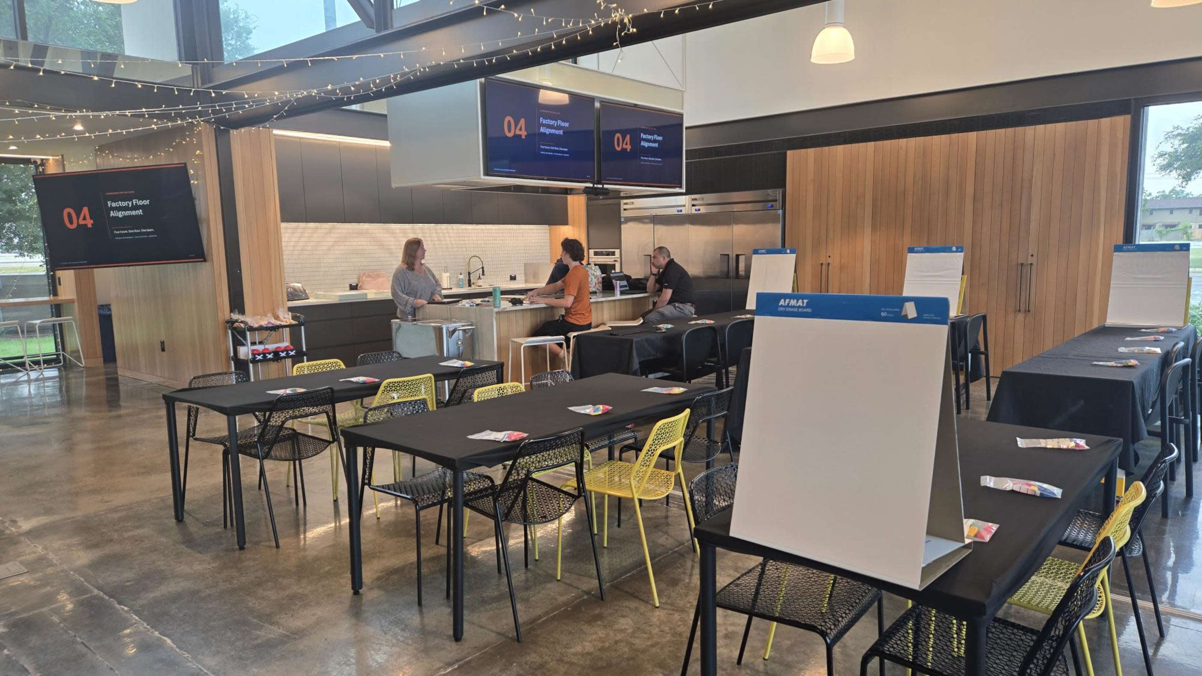The image shows a modern kitchen space with several tables set up for an event, featuring two people at a counter and digital screens displaying information.