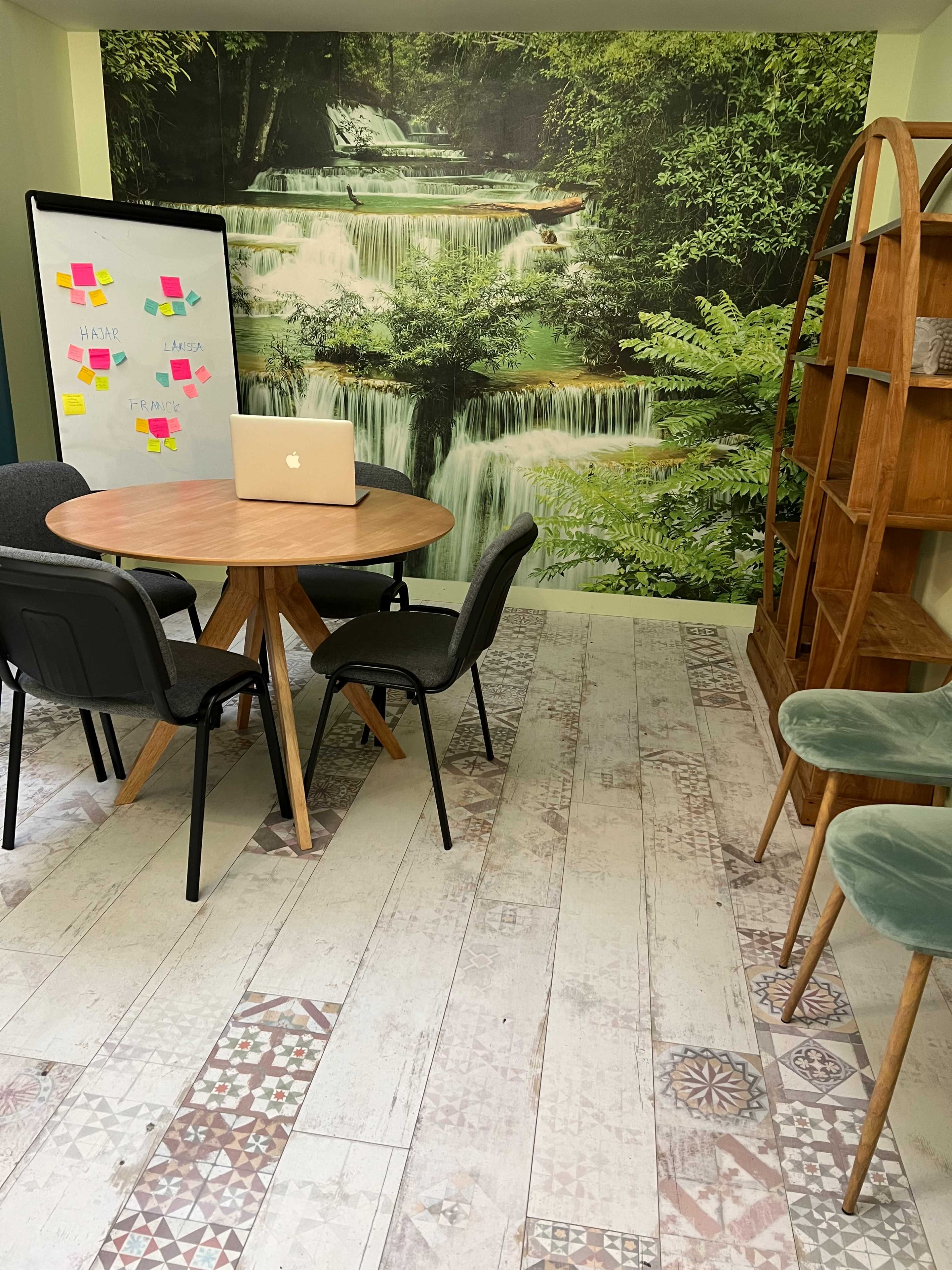 A small meeting area featuring a round table with chairs, a laptop on the table, a wall mural of a waterfall, and a wooden bookshelf against the wall.