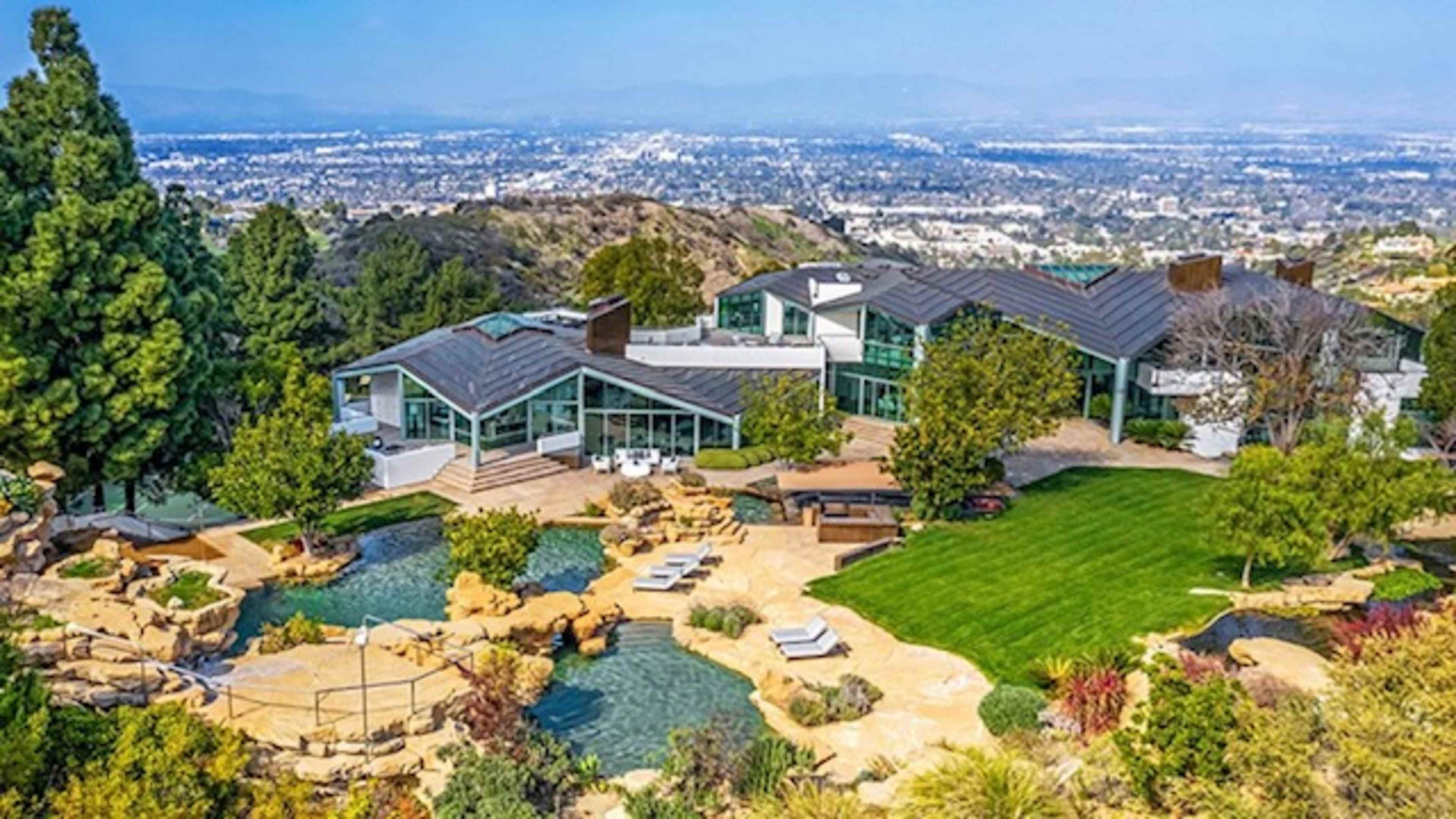 A modern hillside mansion with expansive glass windows, surrounded by landscaped greenery and a swimming pool overlooking a valley cityscape.