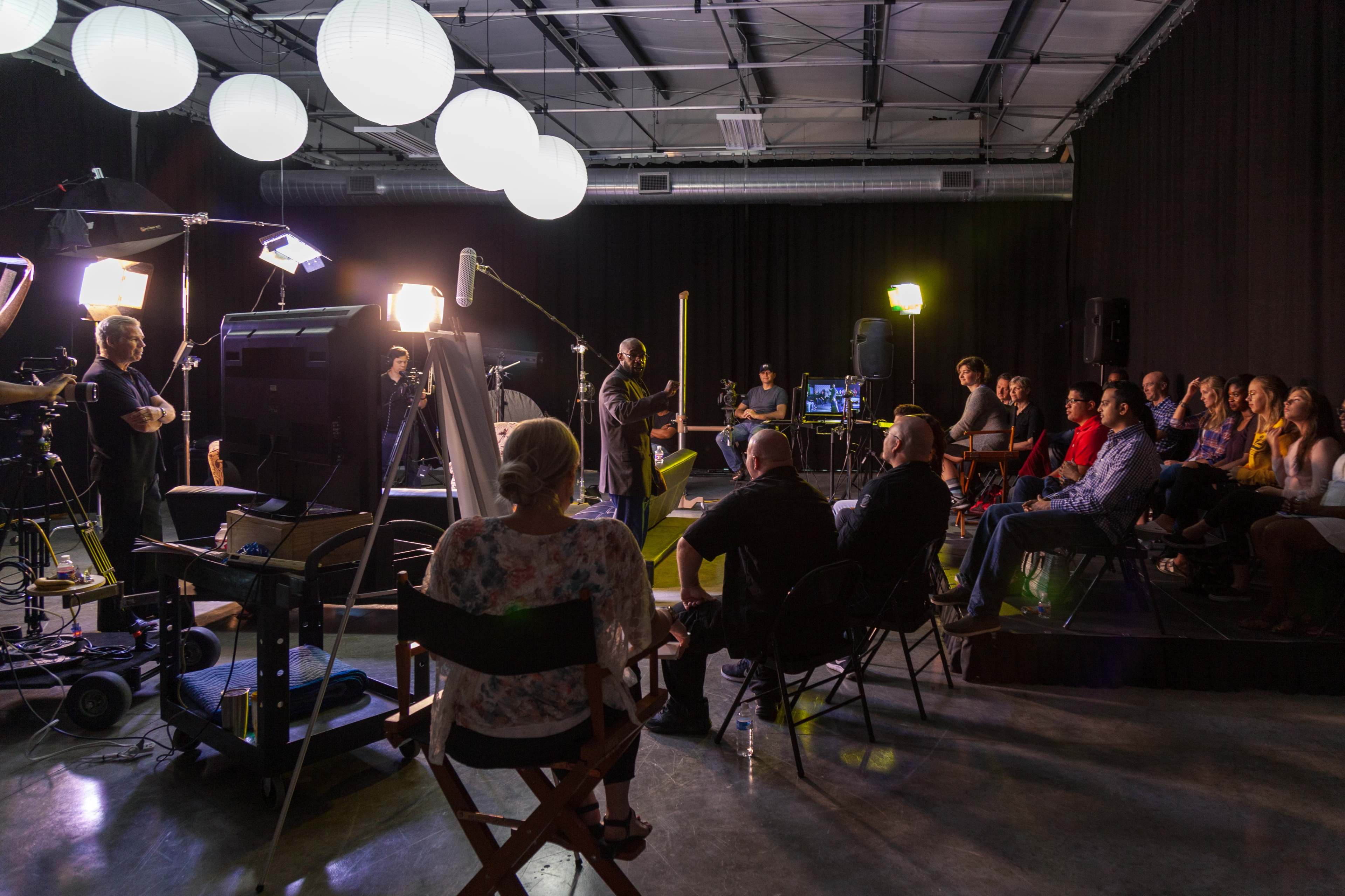 A group of people sits in chairs facing a presenter in a well-lit studio equipped with various cameras and lighting equipment.