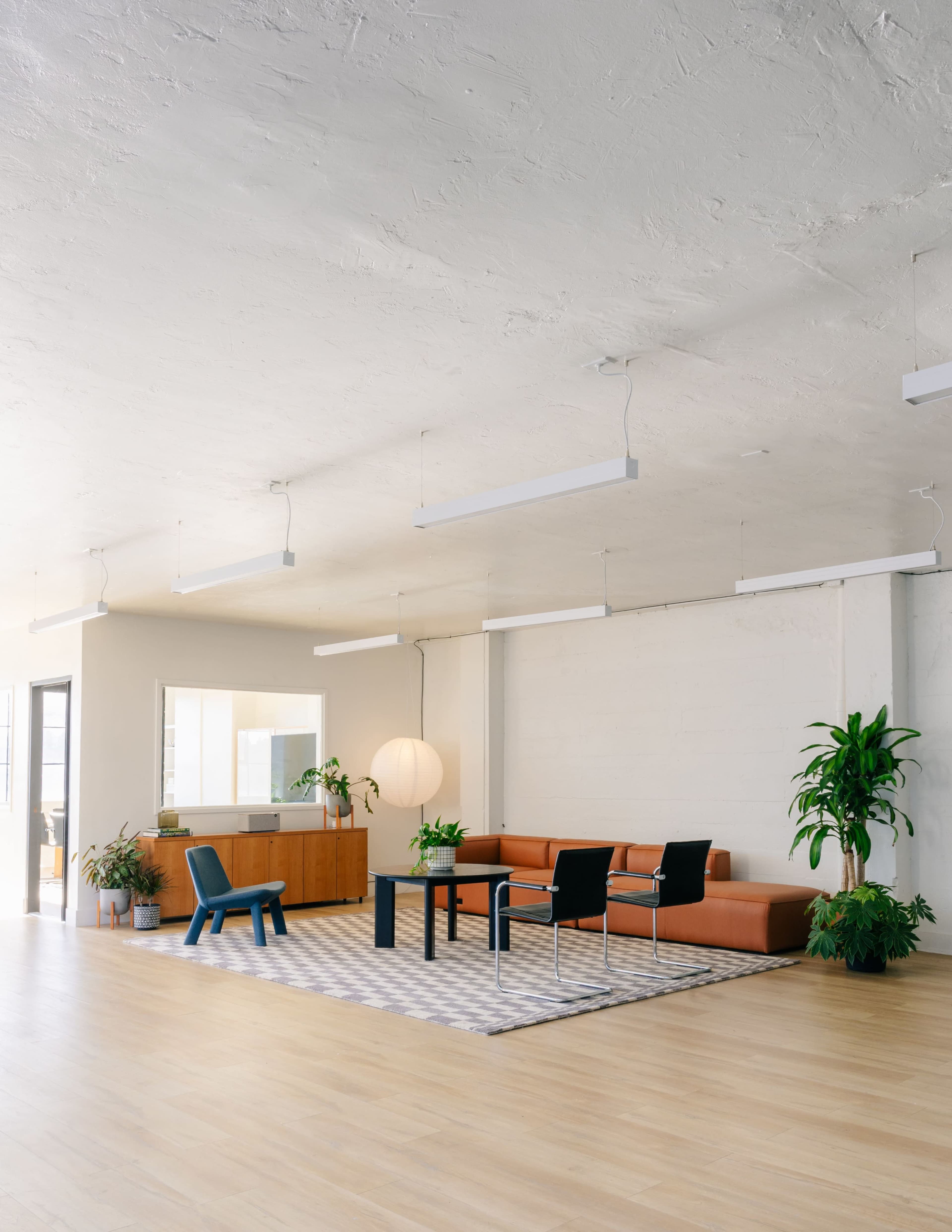 A modern office space features an orange couch, a rectangular table surrounded by black chairs, and plants against a light-colored wall with ample natural light.