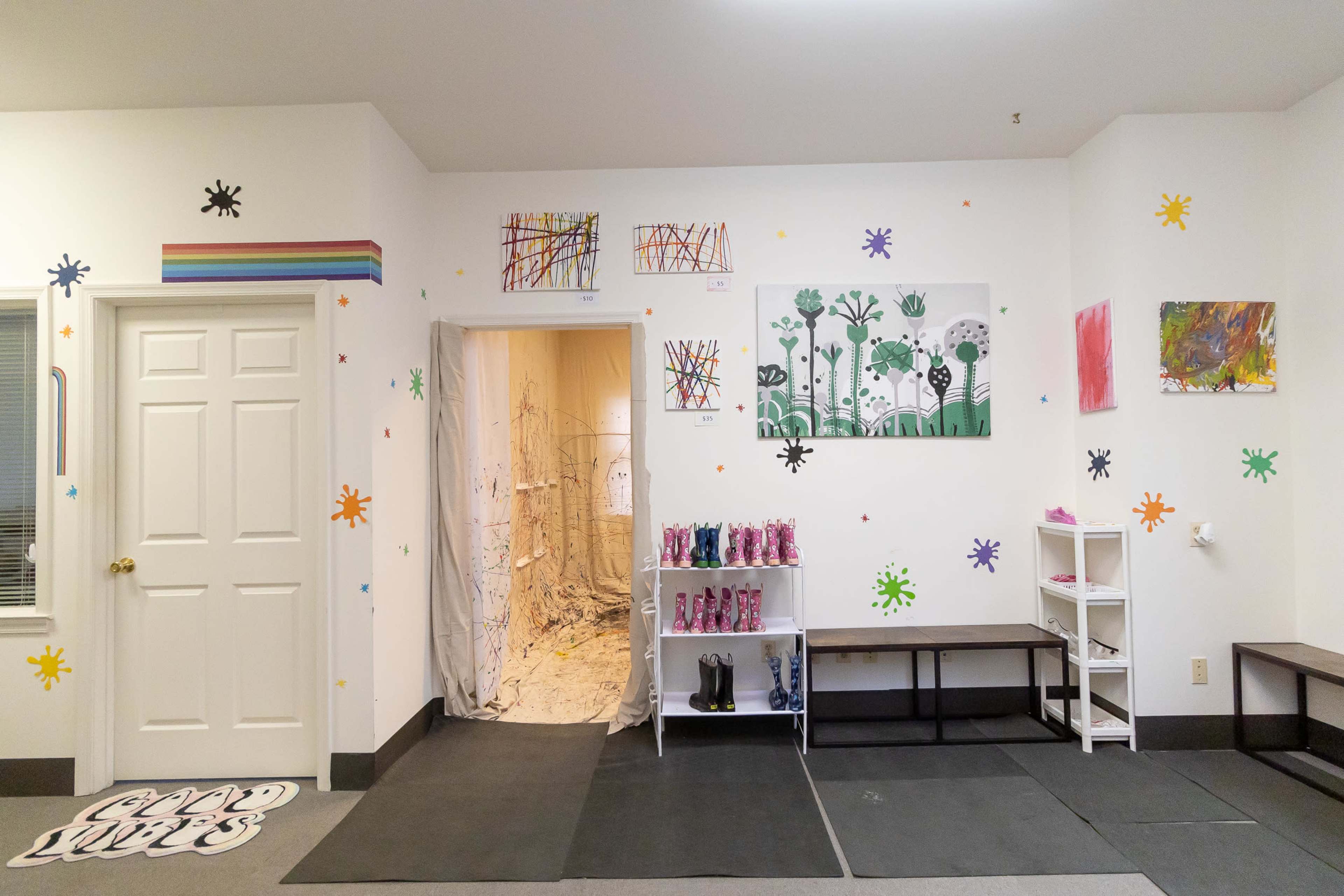 A brightly decorated room features colorful artwork on the walls, a doorway leading to a messy area, and shelves with various items.