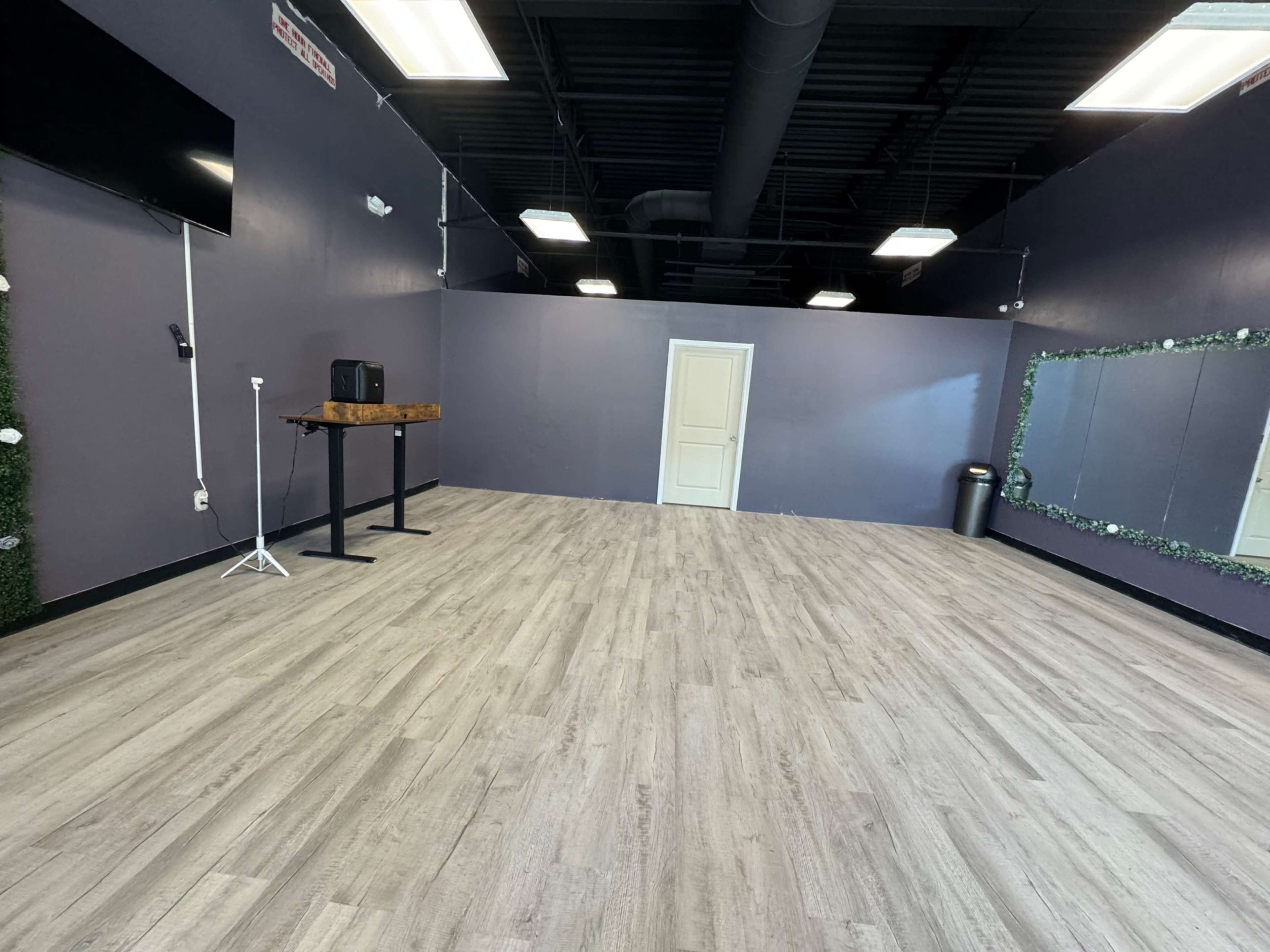The image shows an empty studio space with light wood flooring, a plain purple wall, a full-length mirror, and minimal furnishings.