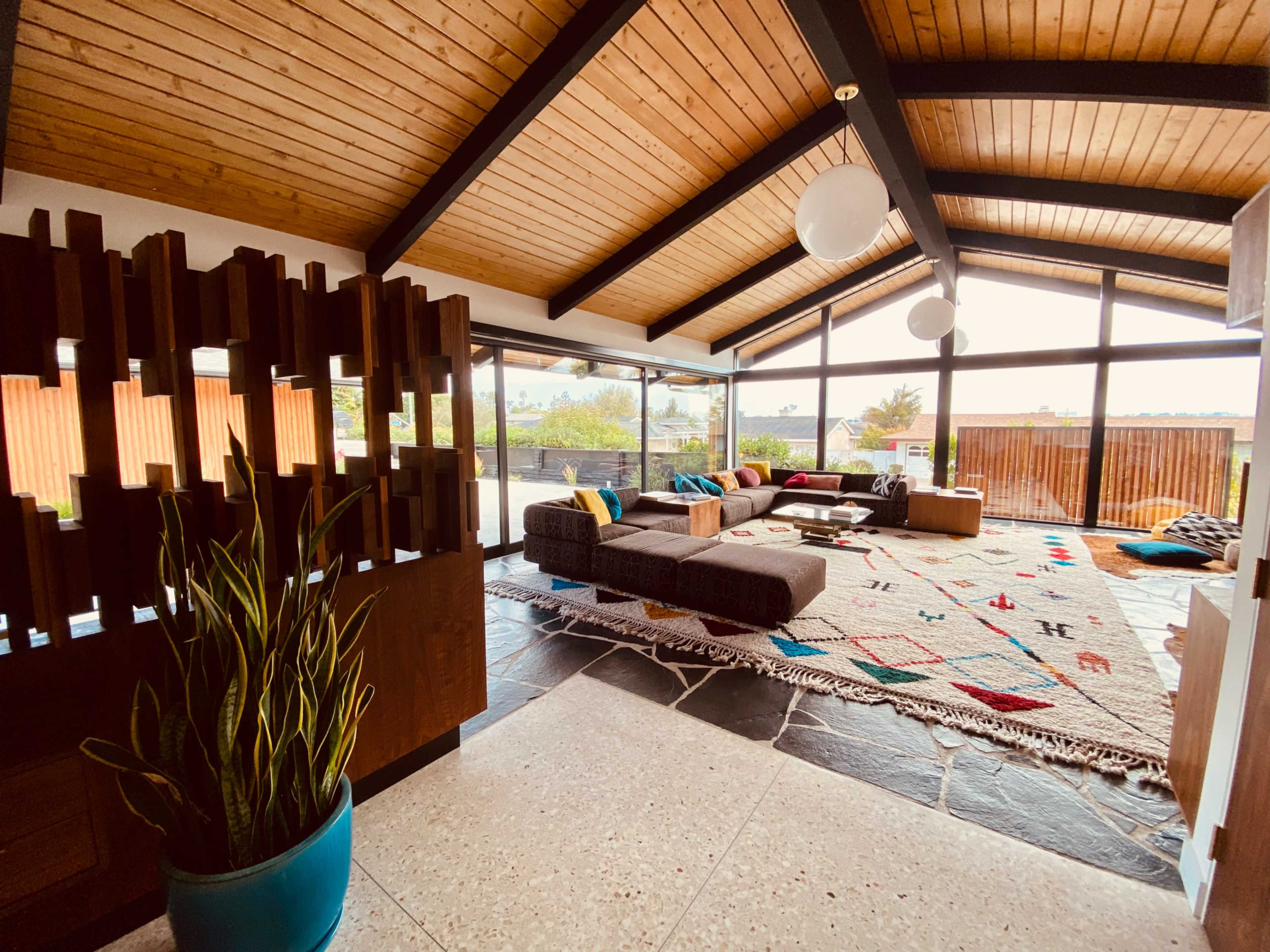 The image shows a modern living room with a high wooden ceiling, large windows, and a comfortable seating area arranged around a patterned rug.