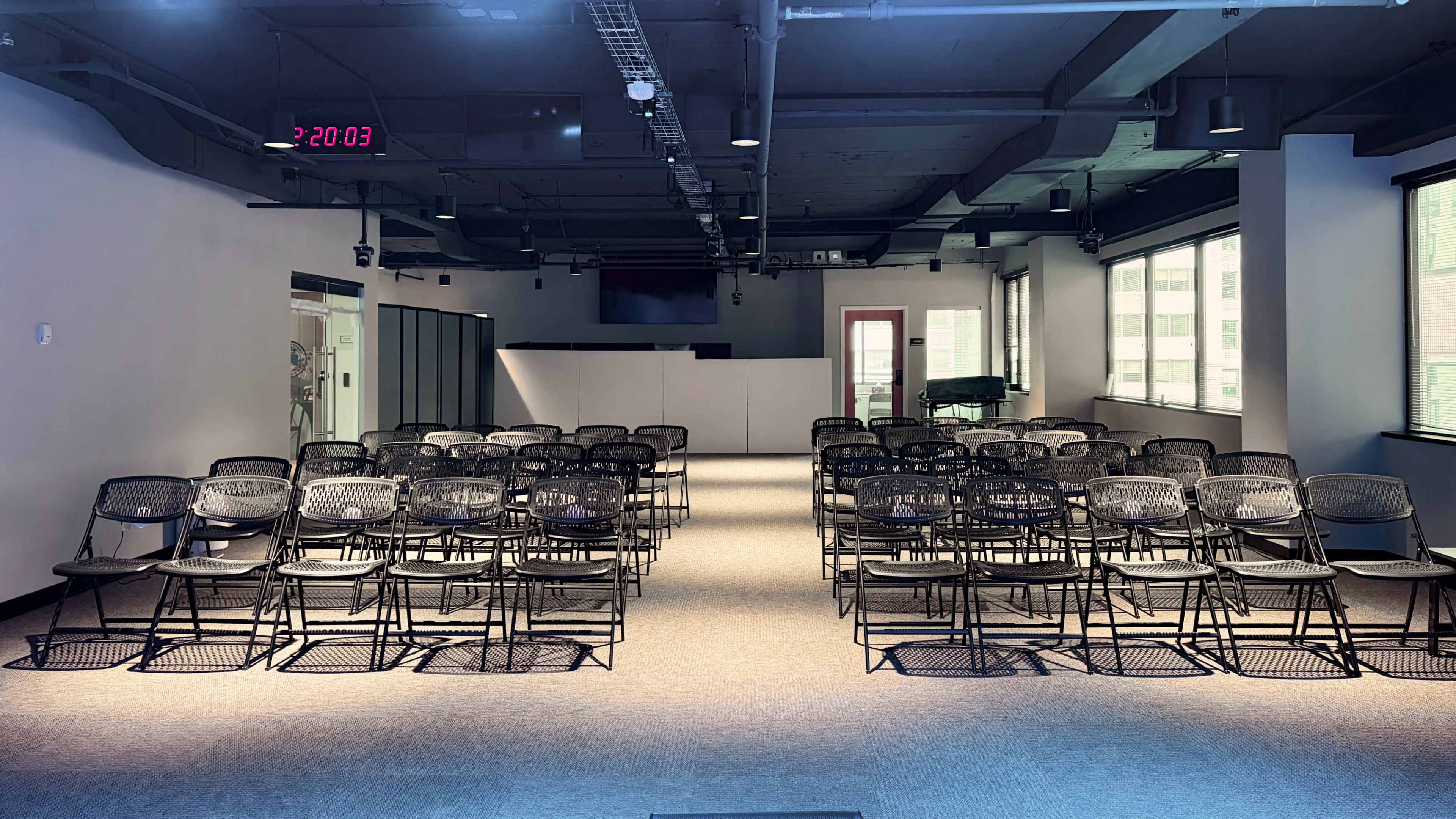 The image shows a spacious, well-lit room arranged with rows of black chairs facing a blank wall and a digital clock displaying the time.