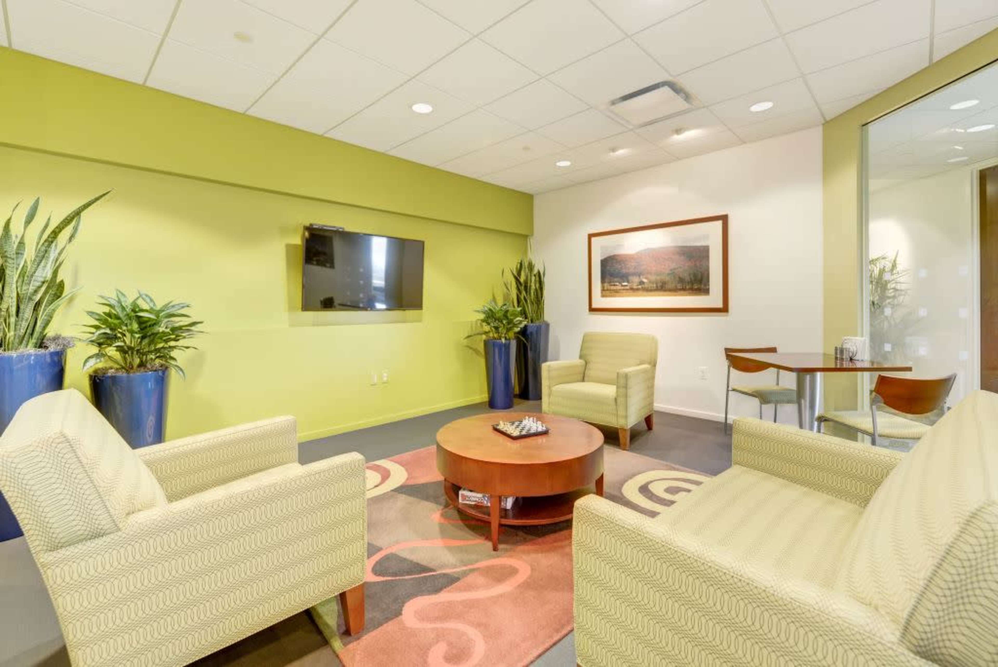 The image shows a modern waiting area with two chairs, a round table, a flat-screen television, and potted plants against a green wall.