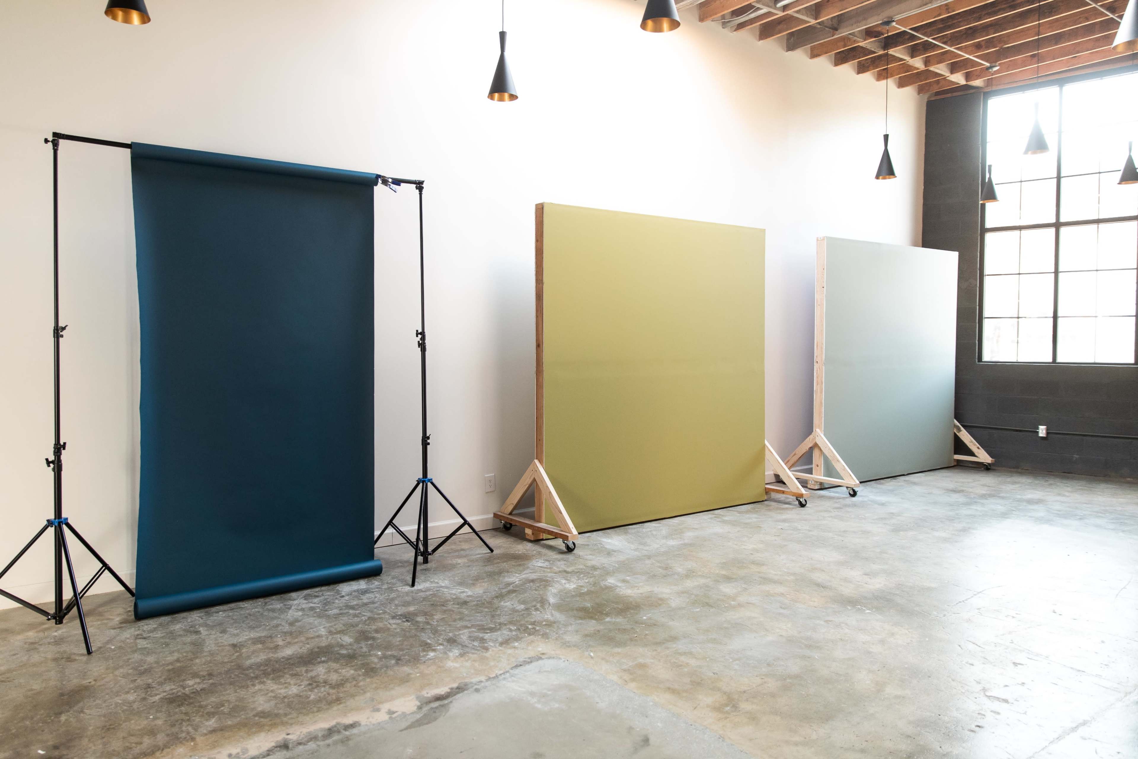The image shows a spacious studio with three different colored backdrops on wooden stands and a backdrop support system, against a plain wall with pendant lighting.