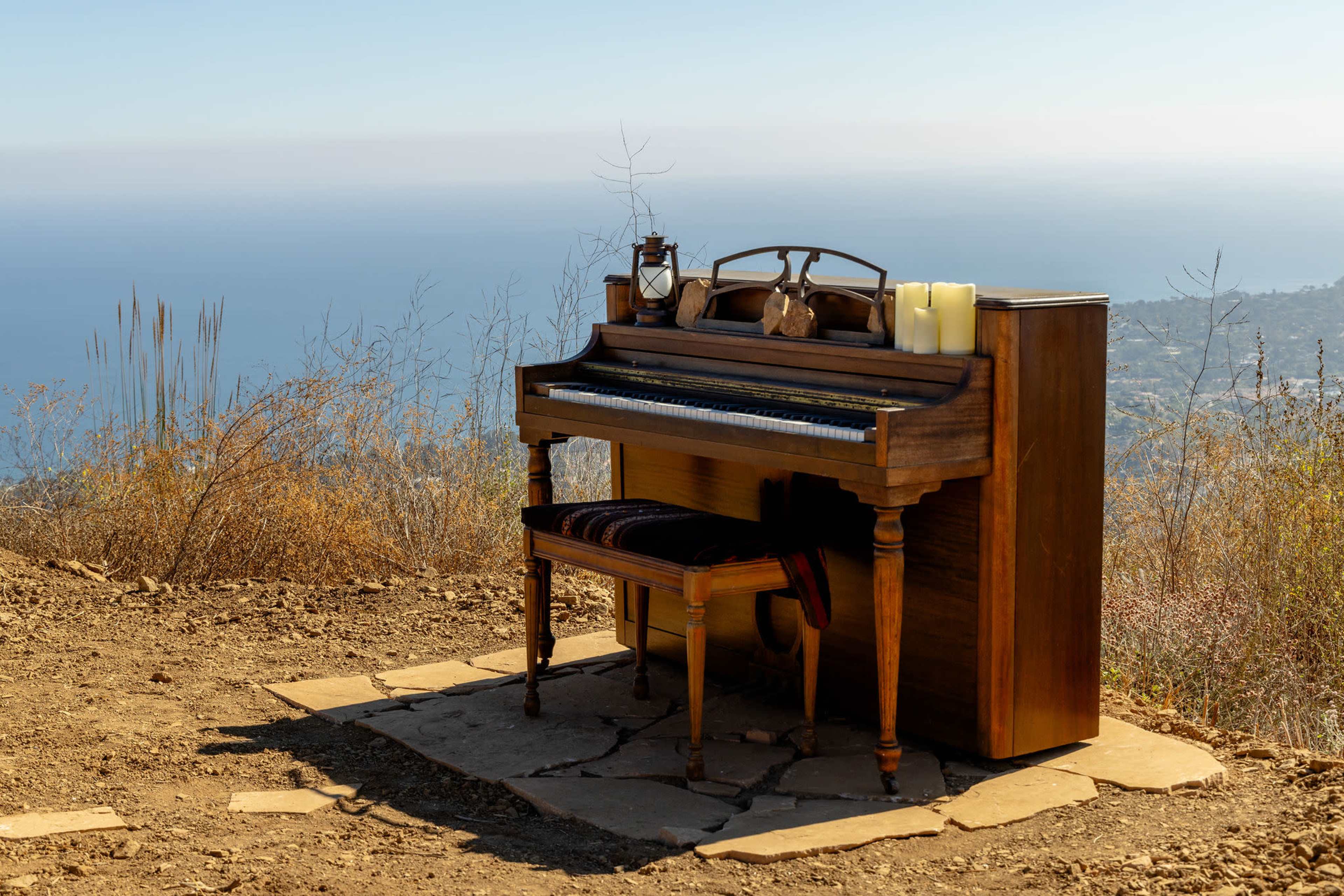 A vintage wooden piano stands on a rocky surface overlooking a vast body of water with a clear sky in the background.