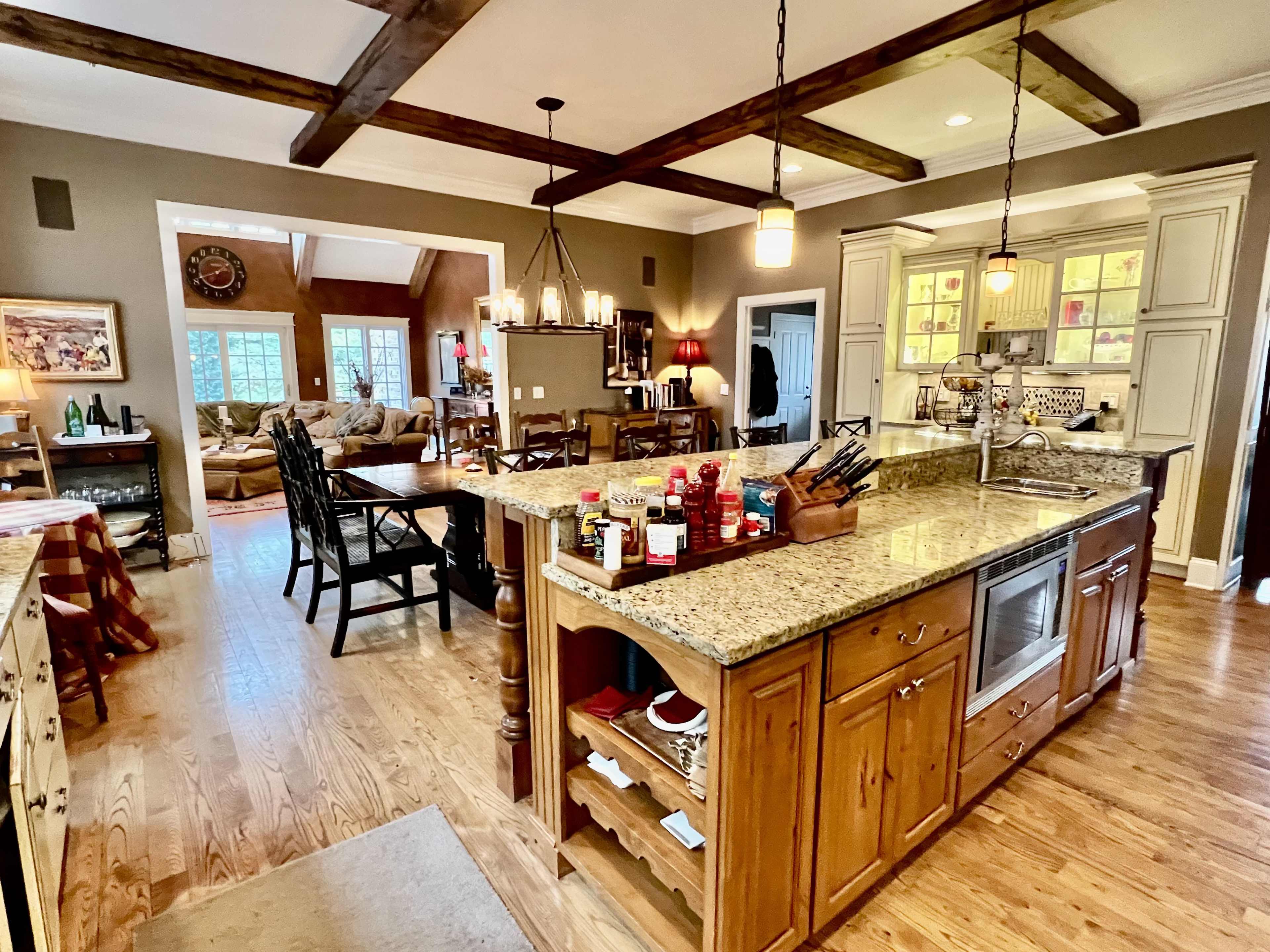 The image shows a spacious kitchen with wooden cabinetry, a central island with a granite countertop, and an open layout leading into a living area.