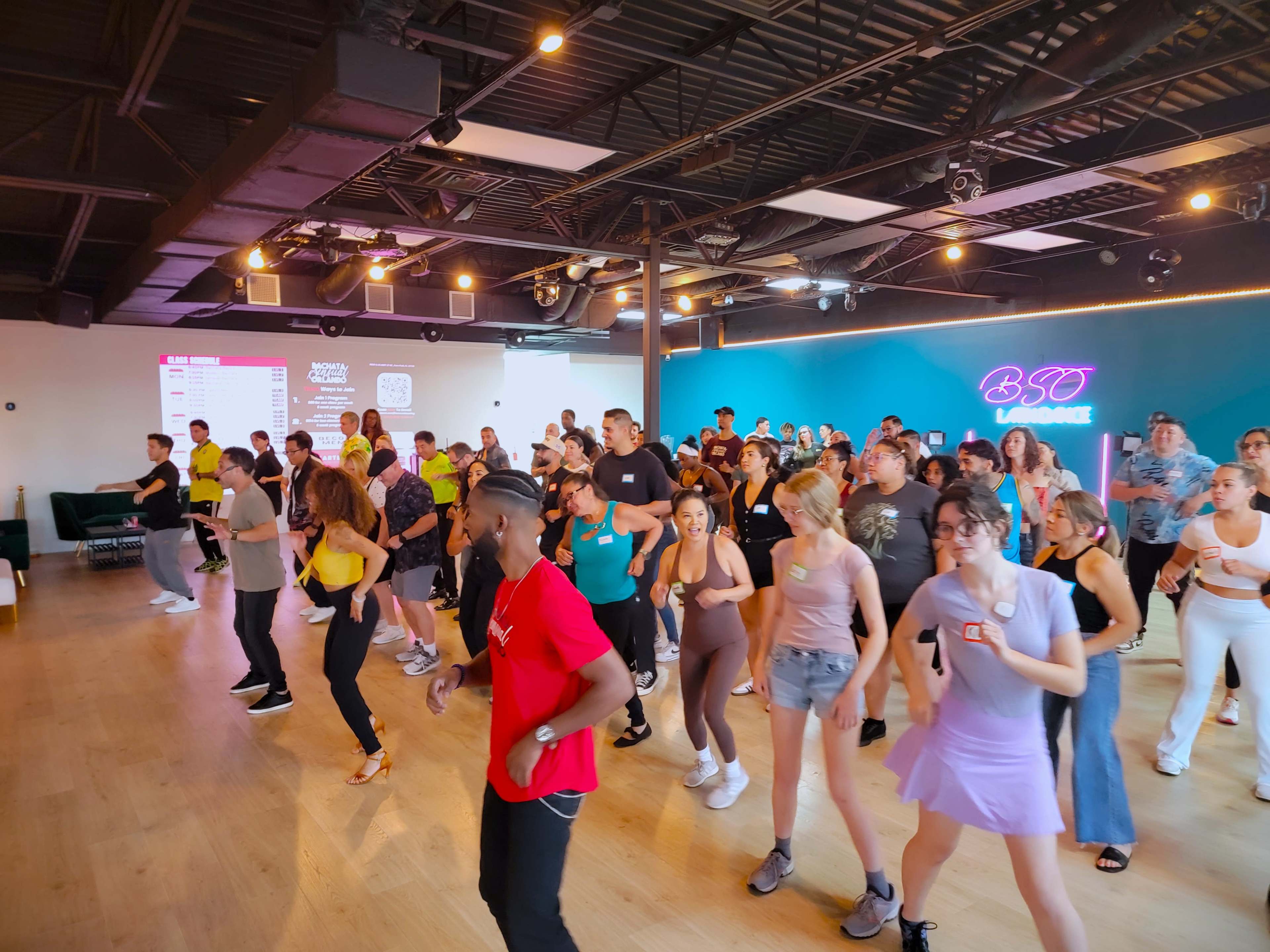 A large group of people is participating in a dance class in a studio with bright lighting and a colorful backdrop.
