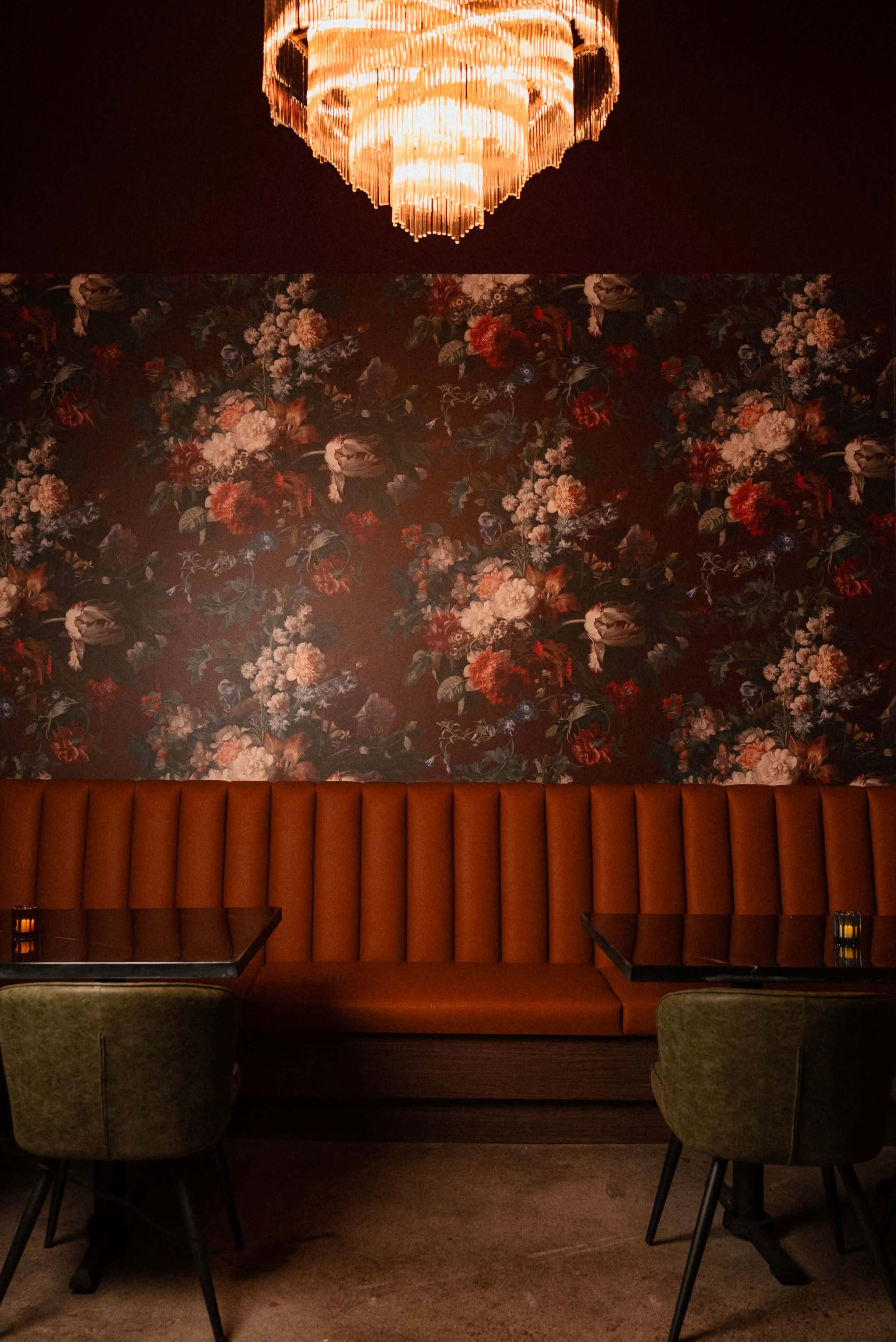 The image displays a dimly lit restaurant interior featuring a floral wallpaper backdrop, an ornate chandelier, and a cozy orange banquette with green chairs around glass-topped tables.