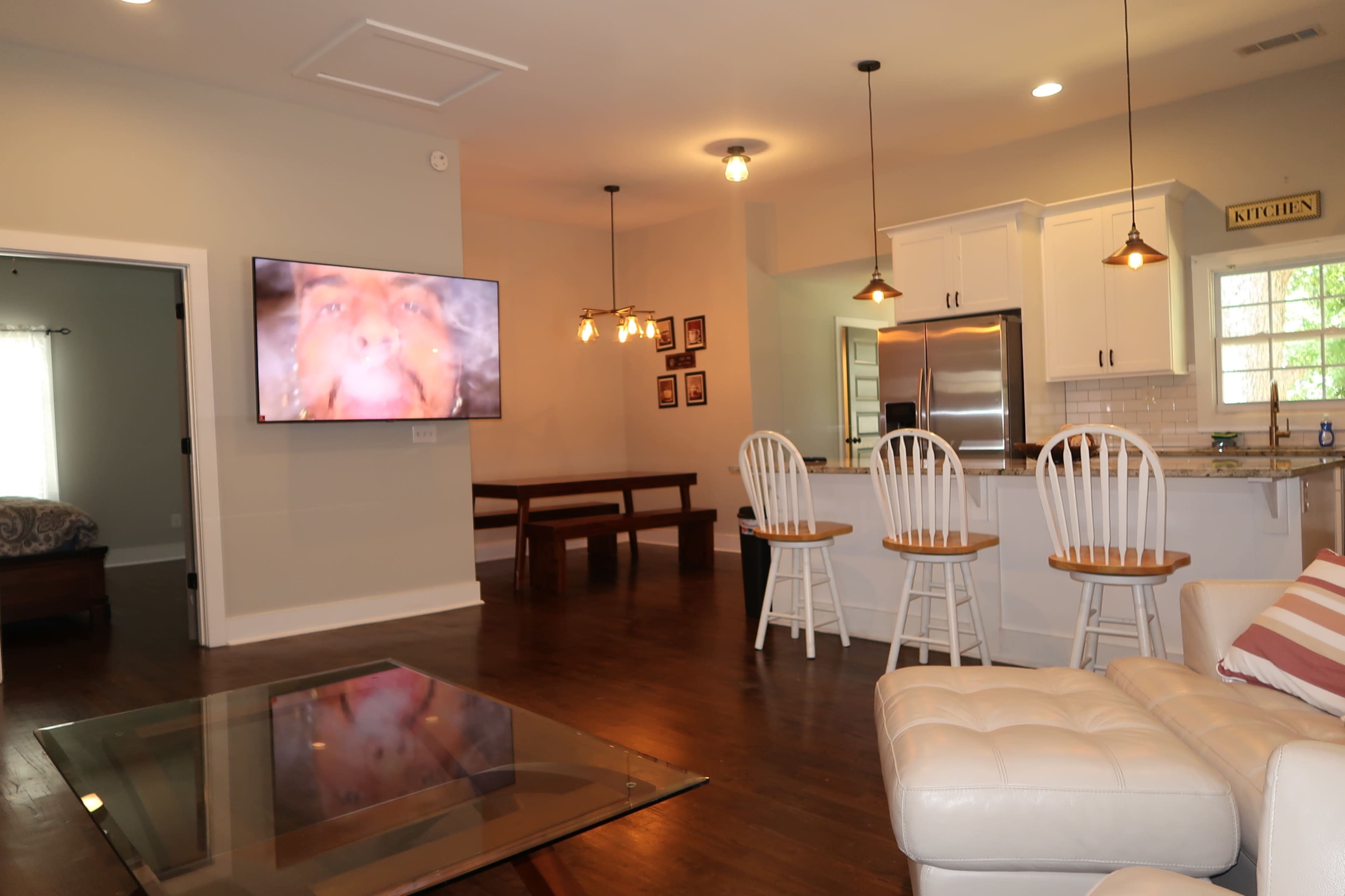 The image shows a modern living room with a large TV mounted on the wall, a glass coffee table, and a kitchen area with white cabinets and bar stools.