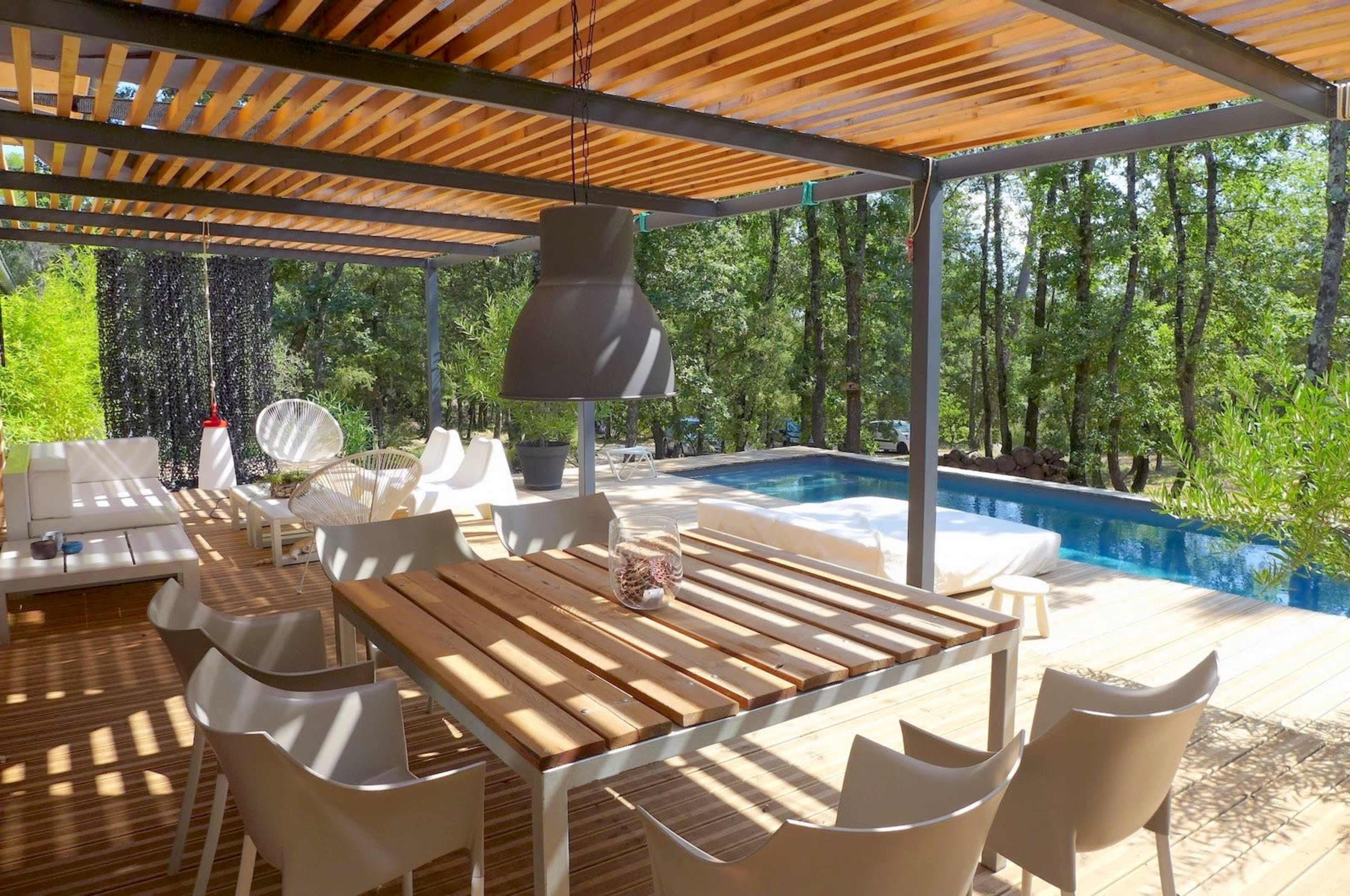 A modern outdoor seating area features a wooden table surrounded by white chairs, with a pool and lush greenery in the background.