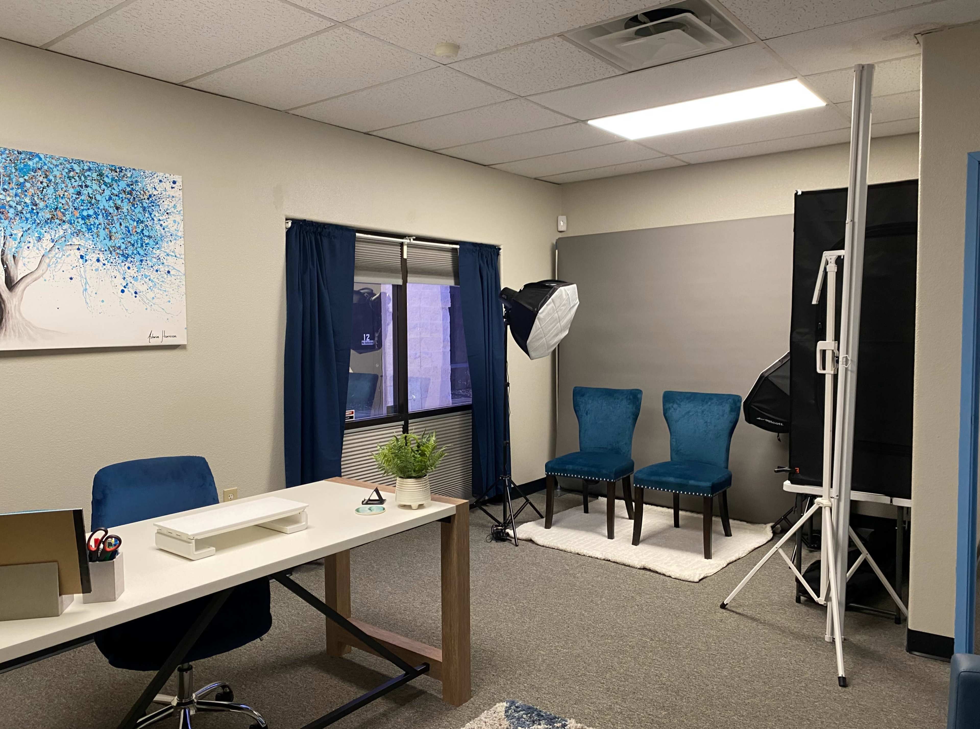 The image depicts a well-organized office space featuring a desk, two blue chairs, a plant, and a backdrop with lighting equipment set up for photography.
