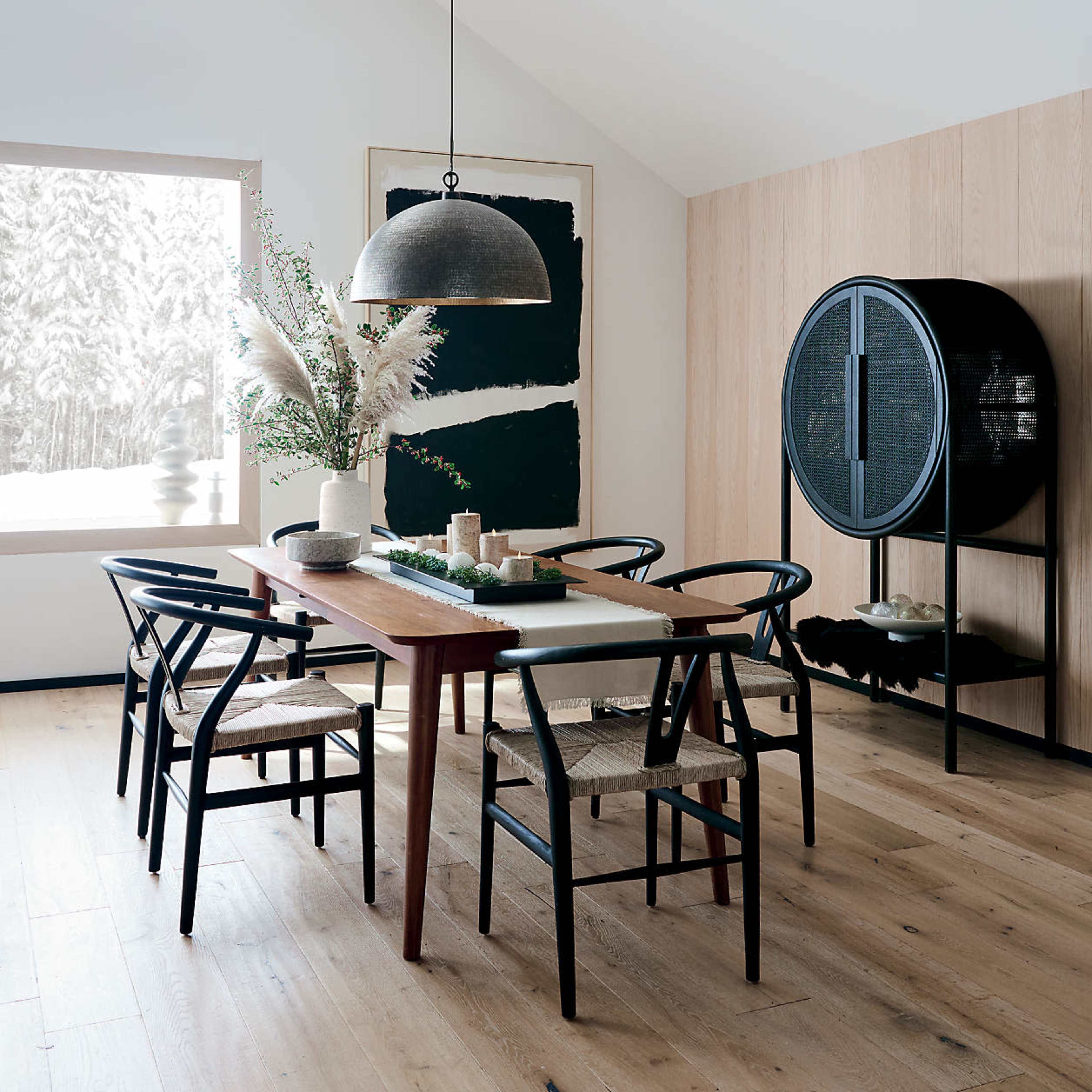 A modern dining room features a wooden table surrounded by black chairs and a decorative cabinet, with a large window showcasing a snowy landscape.