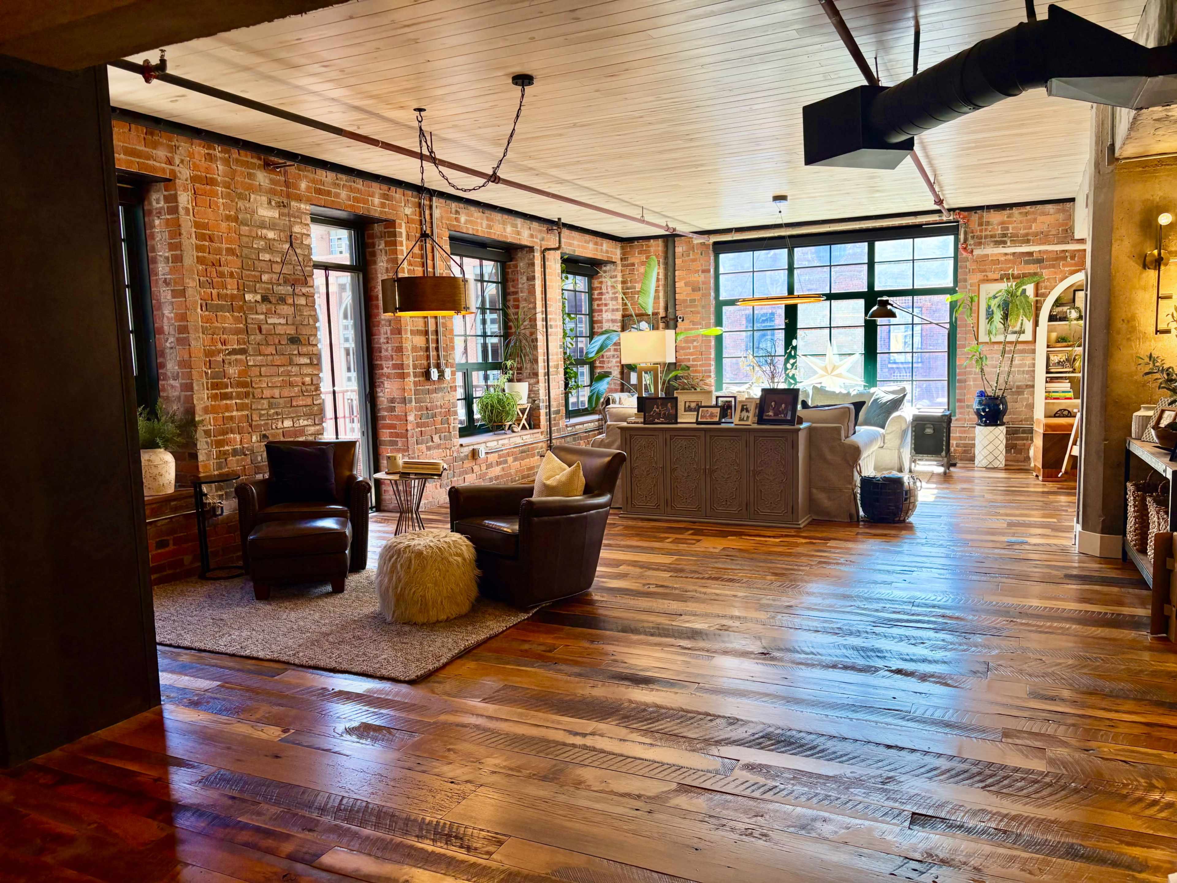 The image shows a spacious, modern living area featuring exposed brick walls, hardwood floors, and large windows that allow natural light to fill the space.