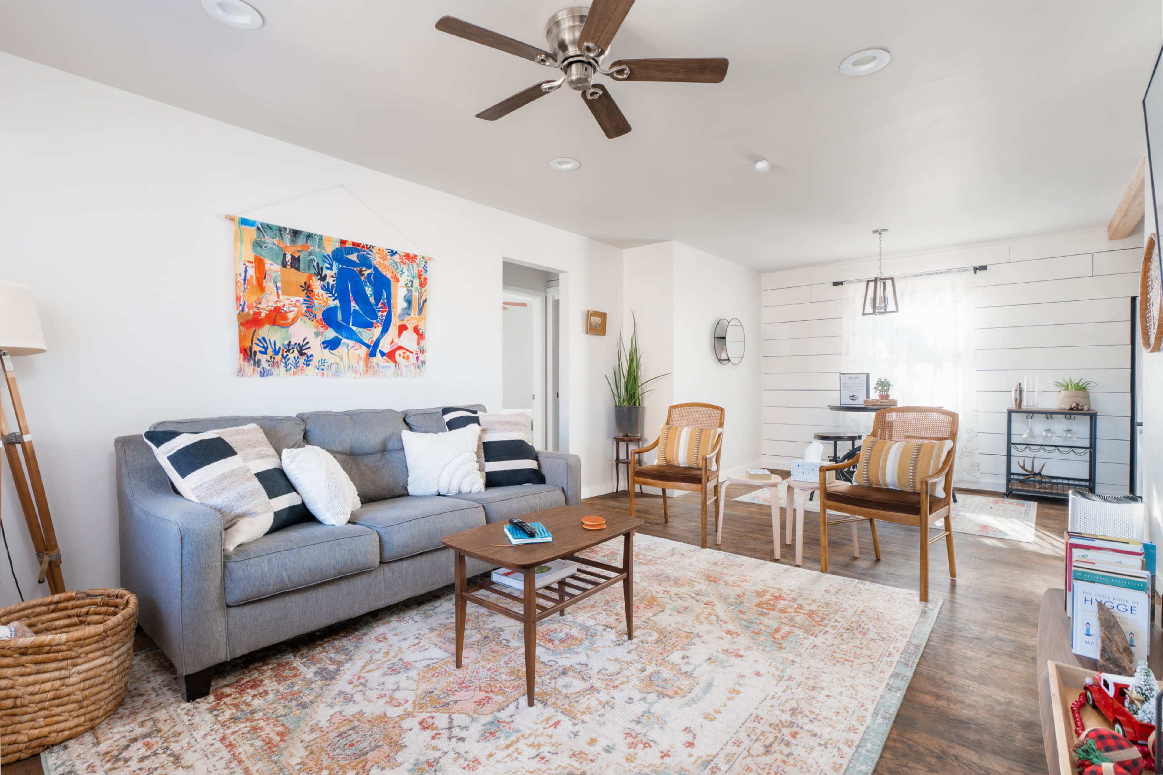 A living room features a gray sofa, two wooden chairs, a small coffee table, and a patterned rug, with artwork hanging on the wall and a plant in the corner.