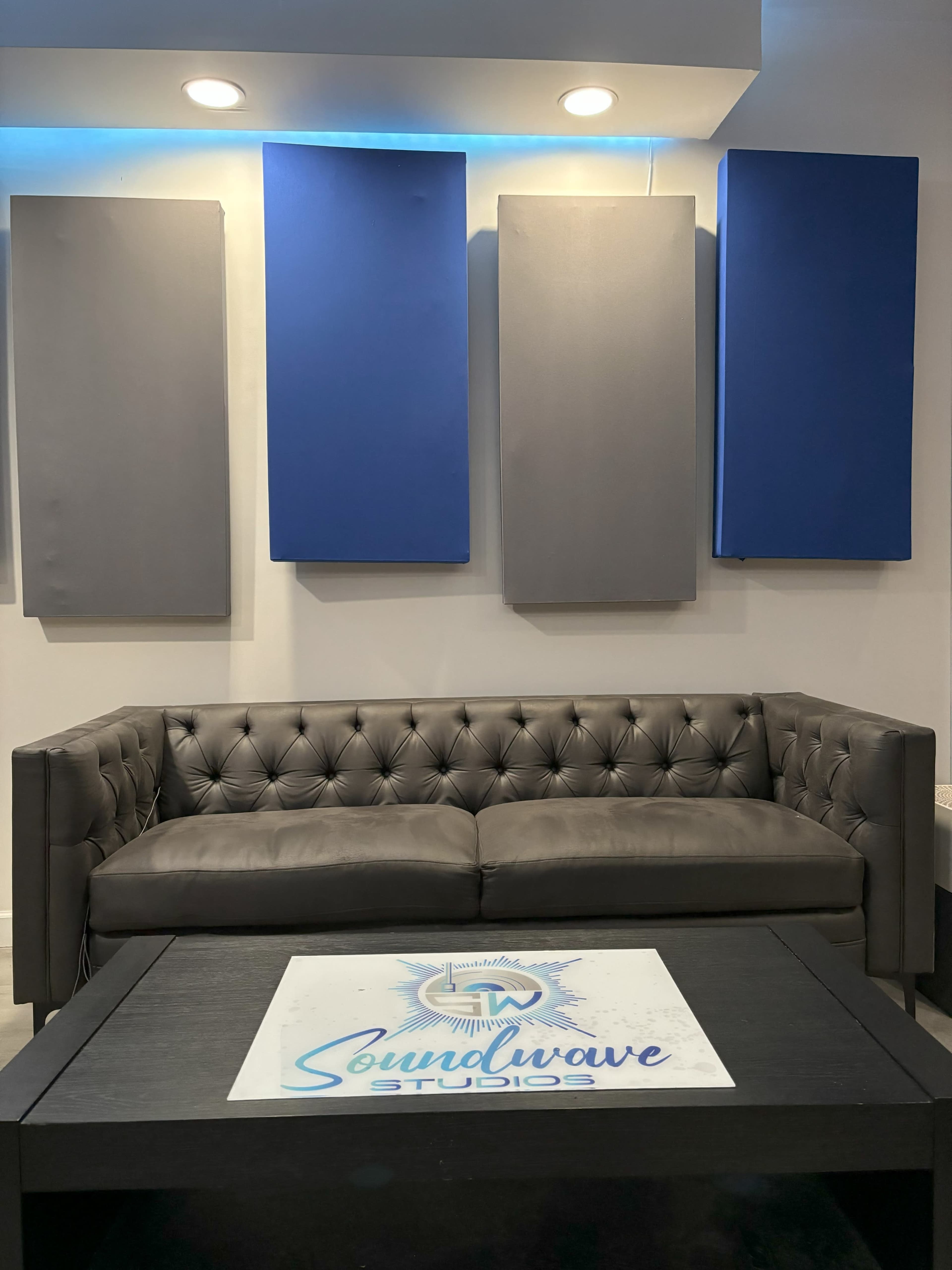 The image shows a gray tufted couch in front of four wall-mounted soundproofing panels, with a coffee table featuring a logo for "Soundwave Studios."