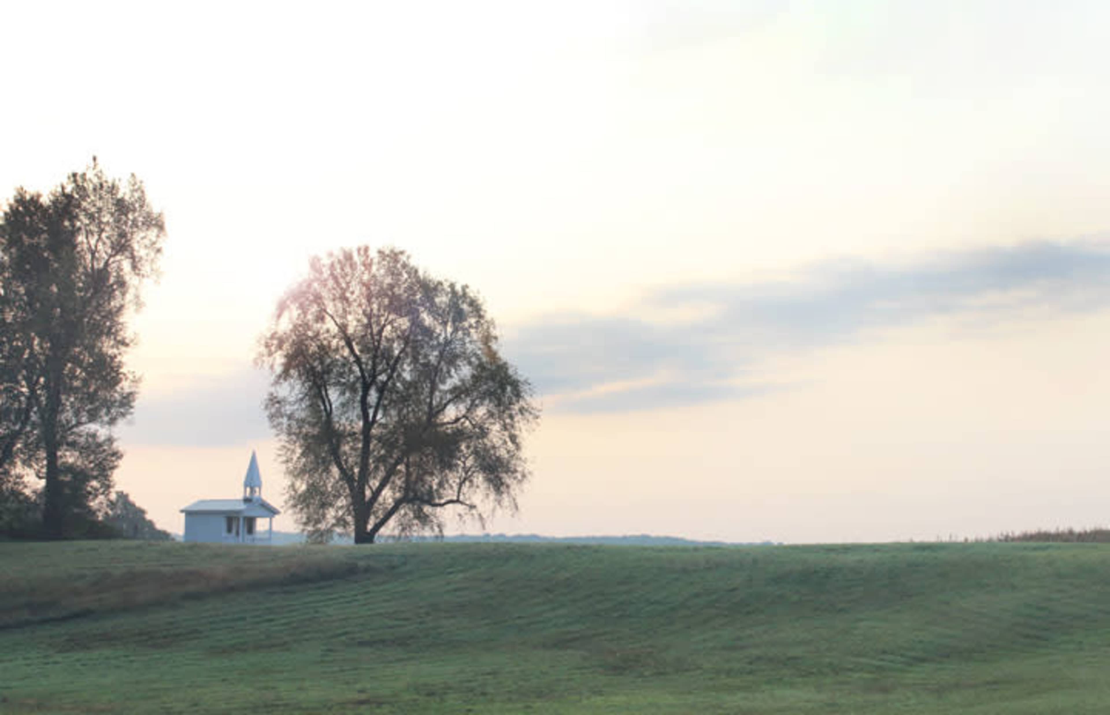 A small white church with a steeple sits near the edge of a grassy field, surrounded by trees under a cloudy sky.