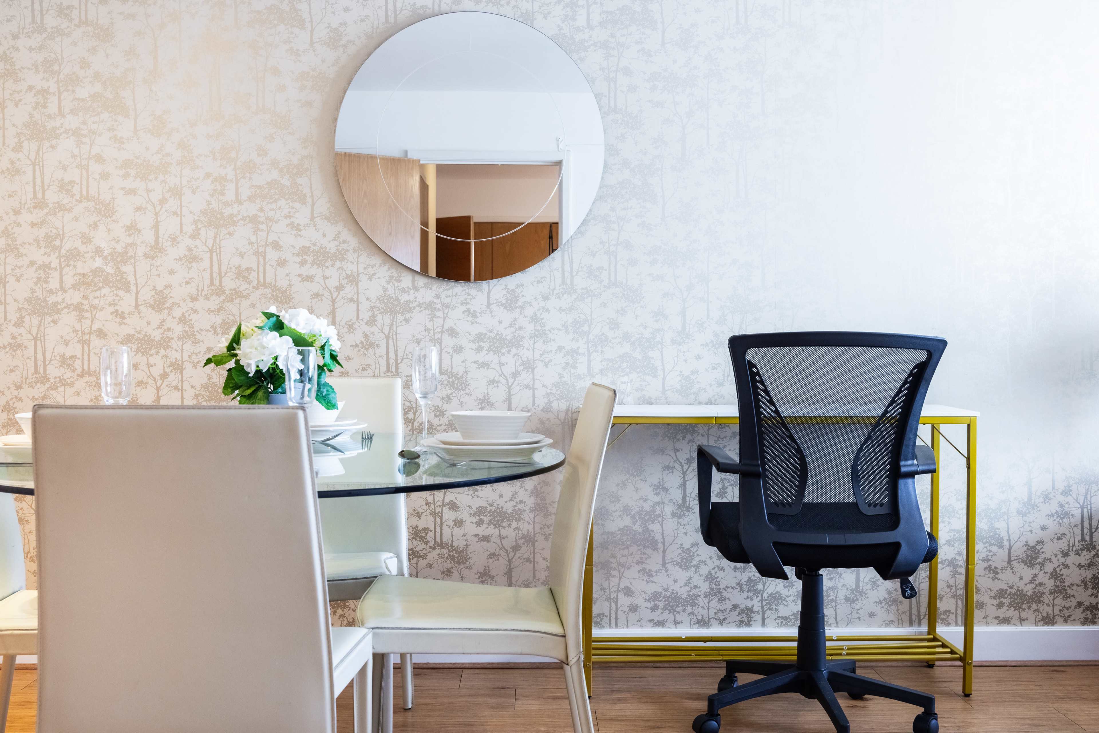 A round glass dining table with white chairs is set next to a mirror and a workspace featuring a black office chair against a patterned wall.