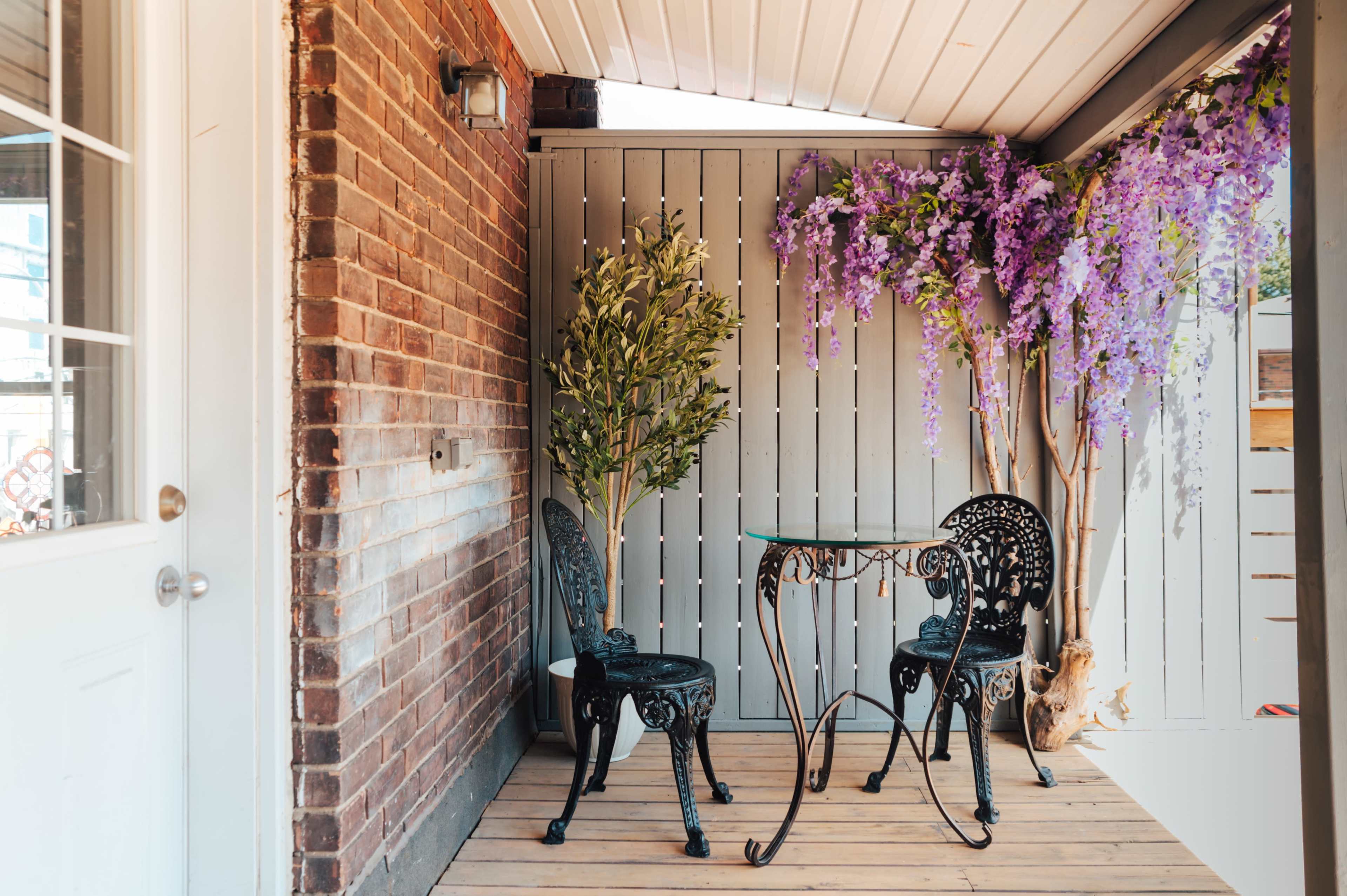 A small porch features a glass table and two black chairs, surrounded by a potted plant and purple wisteria decor along the walls.