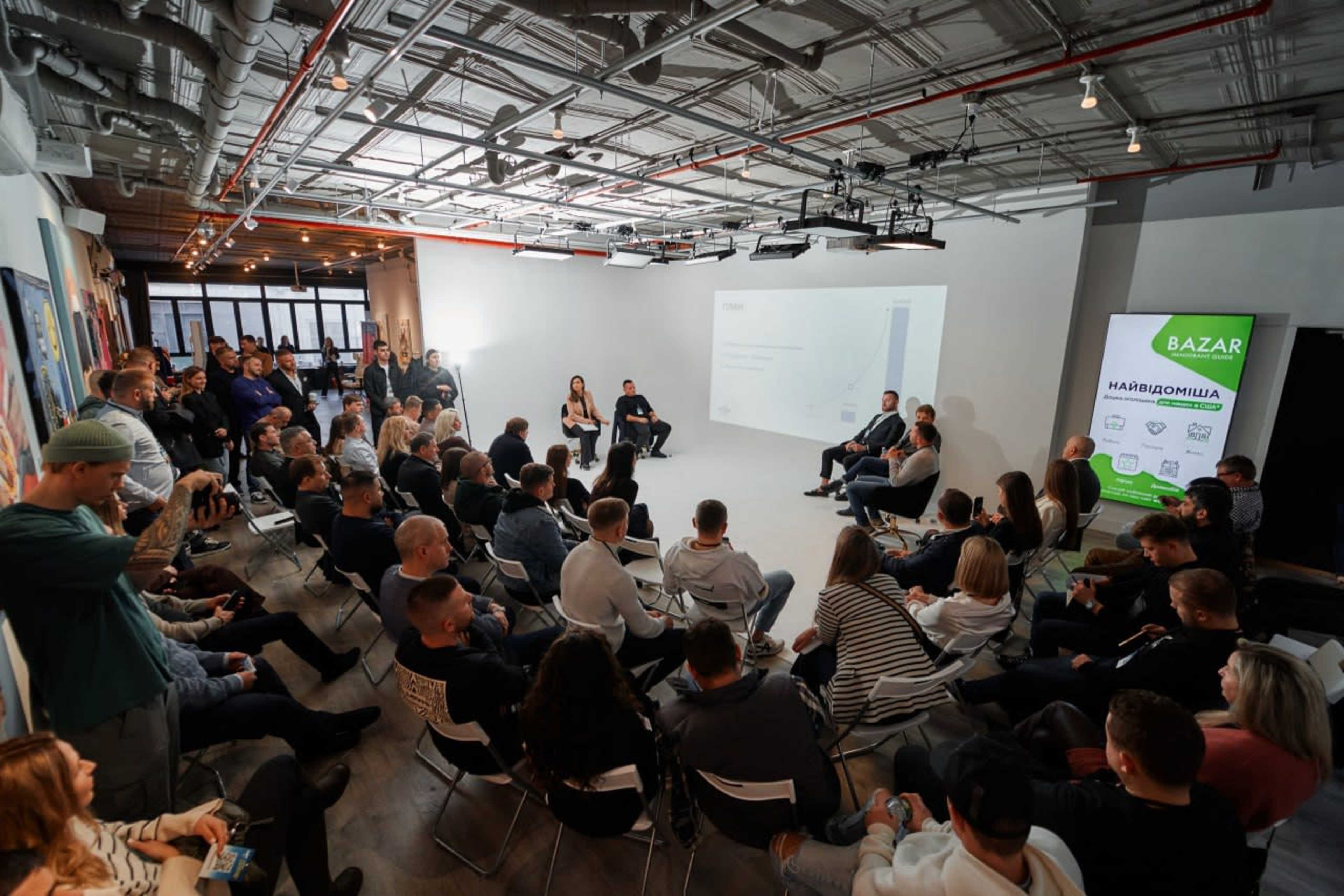 A group of people sits in a well-lit room arranged for a presentation, facing a panel of speakers in front of a white screen.