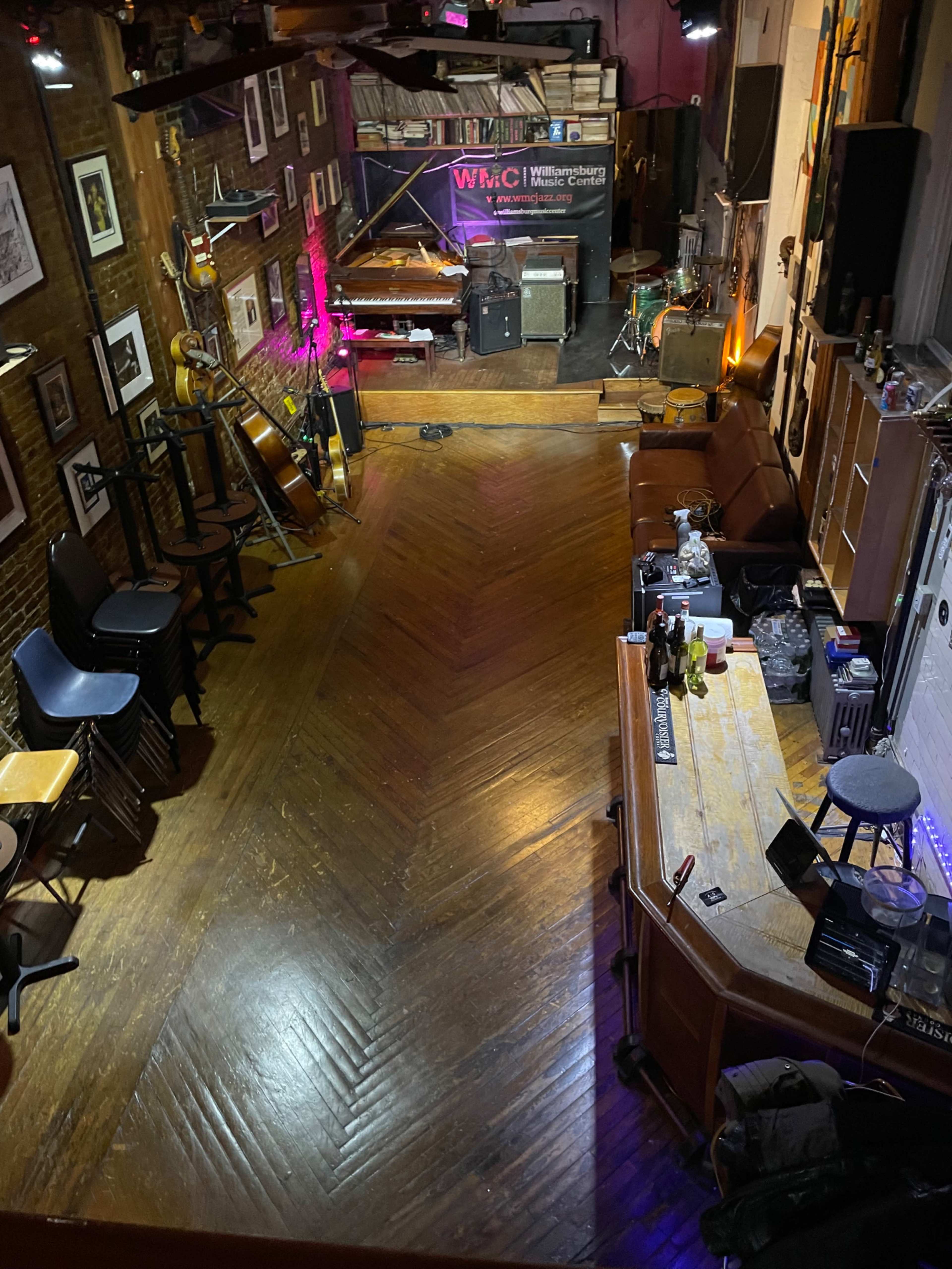 The image shows a small live music venue featuring a wooden floor, a stage with instruments, and a bar area with seating.