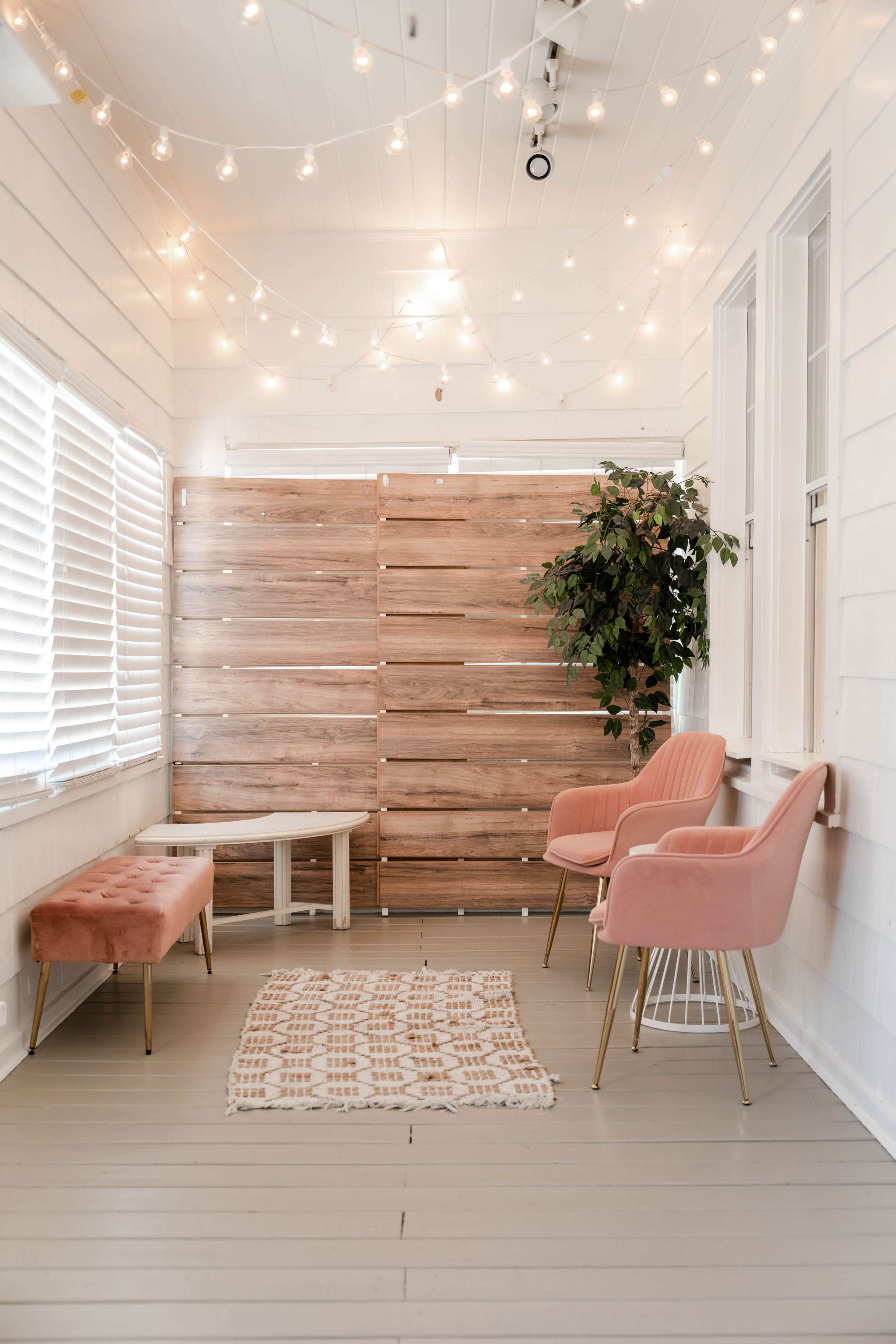 A cozy room features two pink chairs, a small table, and a wooden privacy wall, illuminated by string lights.