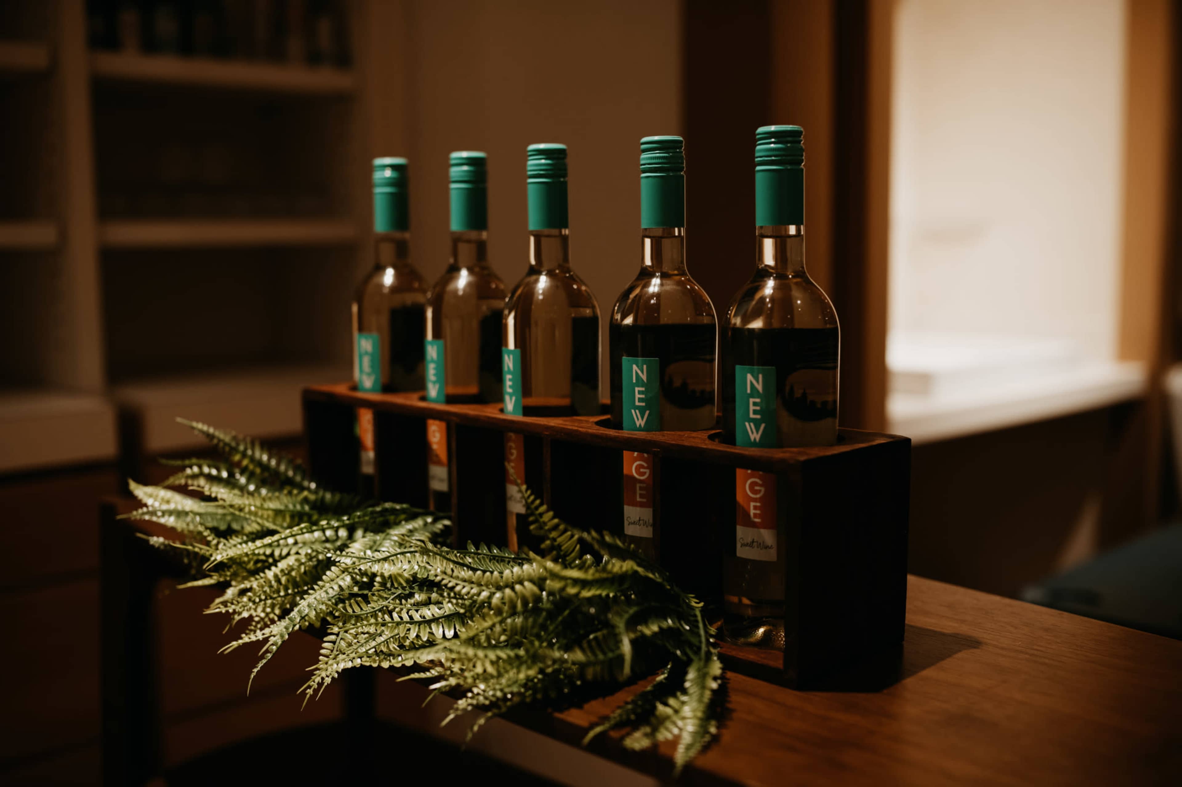 Five bottles of white wine are arranged in a wooden holder on a table, accompanied by a green fern.