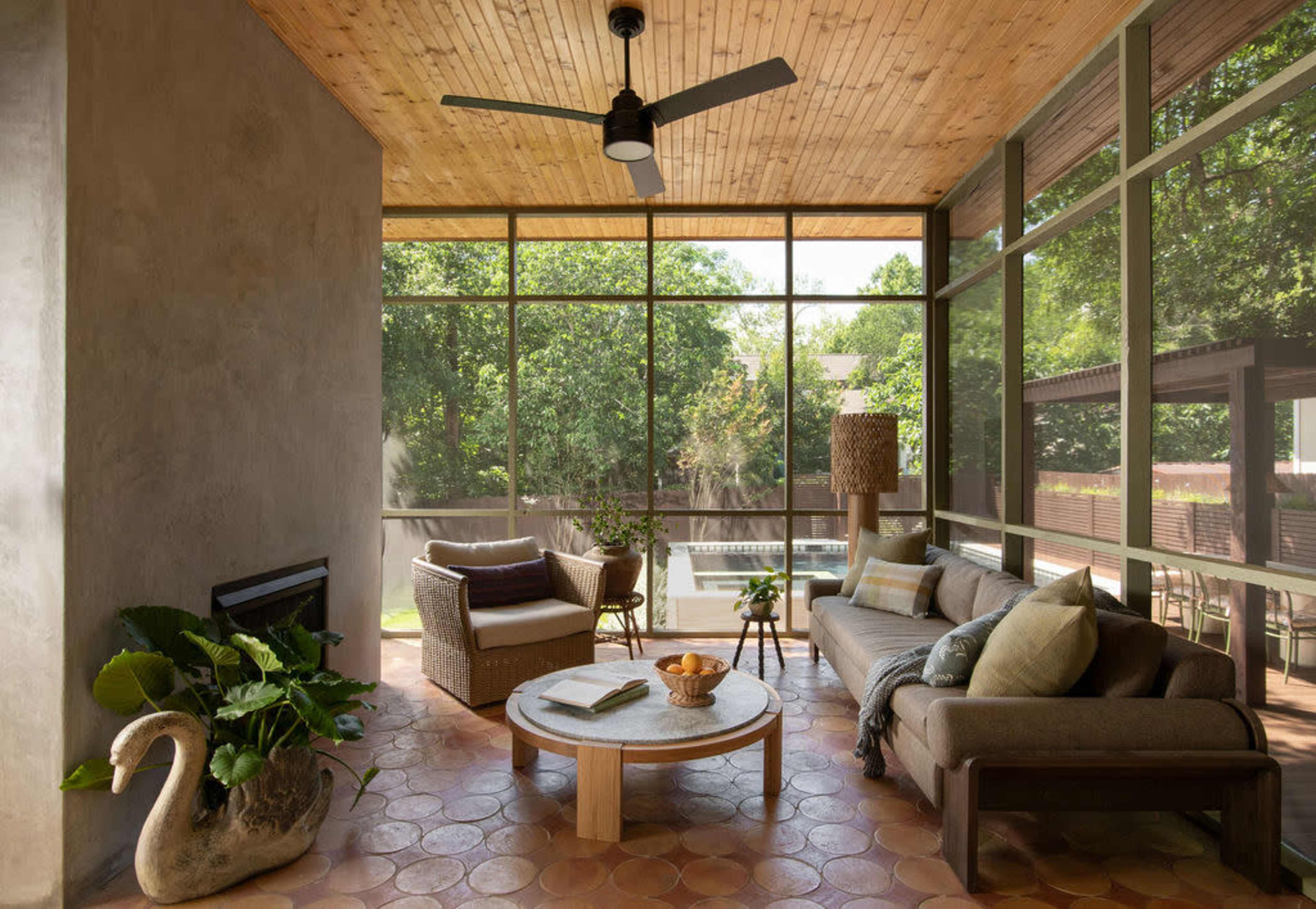 The image shows a modern sunroom with large windows, a ceiling fan, and a seating area featuring a sofa, a chair, and a round coffee table.
