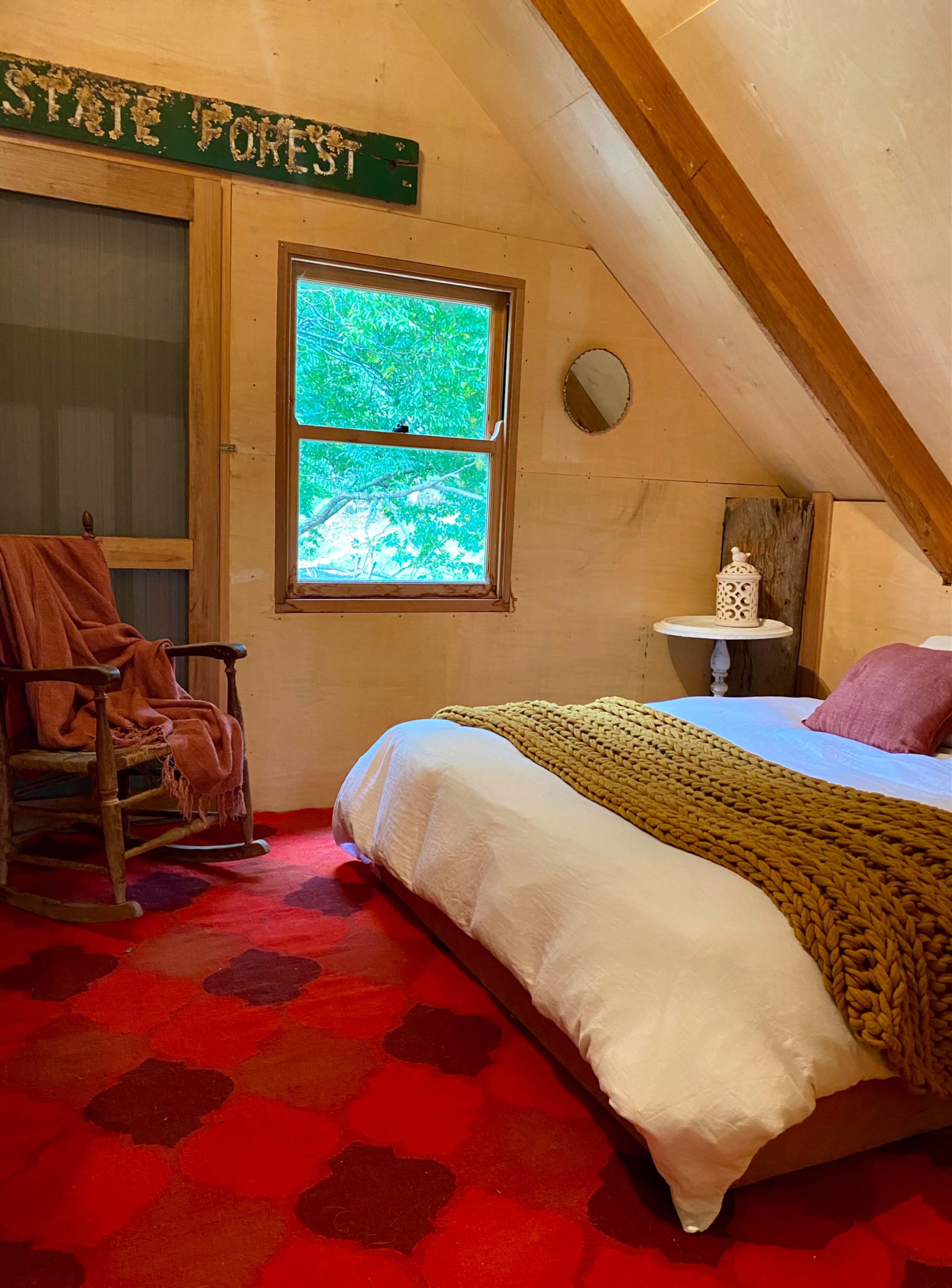 A cozy bedroom features a bed with a knitted blanket, a rocking chair, and a window overlooking a green landscape.