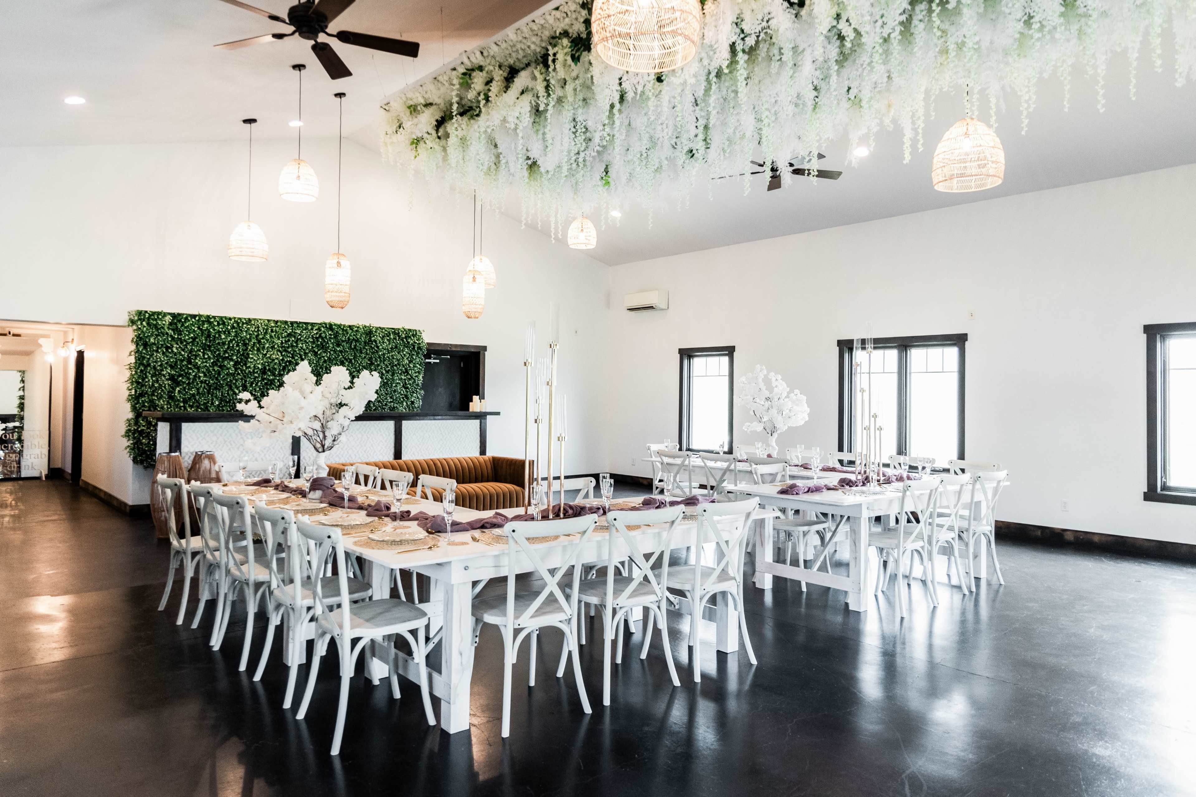 A spacious dining area with white tables set for a meal, surrounded by greenery and pendant lighting.